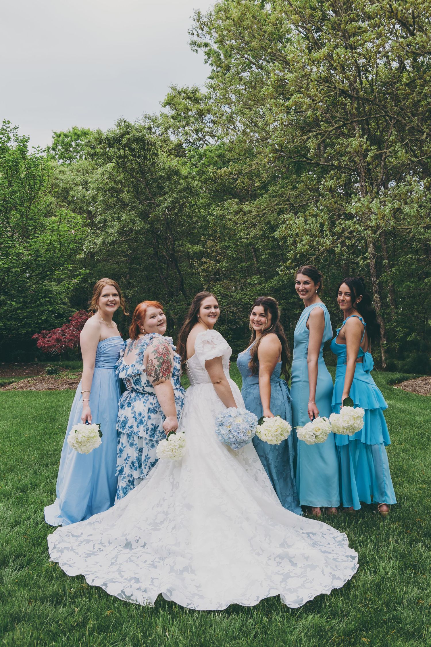 Bridal party in various shades of blue dresses posed together on green grass.