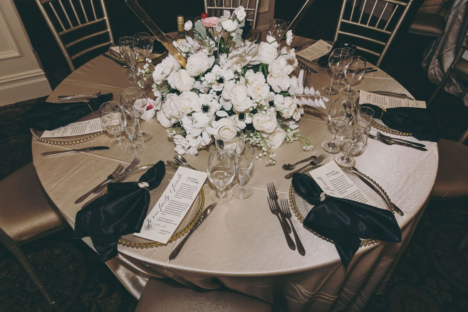 Elegant wedding reception table setting with white floral centerpiece, silver cutlery and place settings on beige tablecloth.