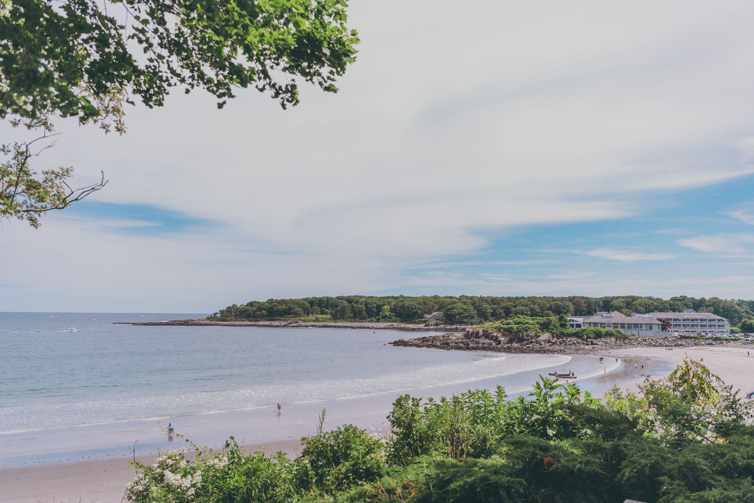 Wide sandy beach stretches along the rugged Maine coastline with boats dotting the harbor in the distance.
