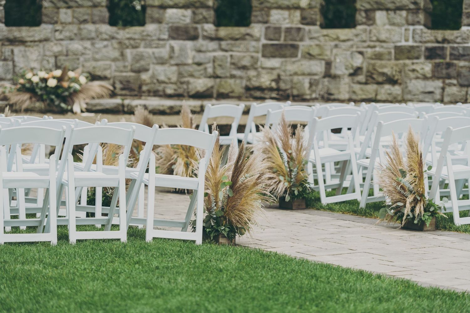 Stone wall backdrop with rows of white folding chairs set up for an outdoor event on grass and paved area.