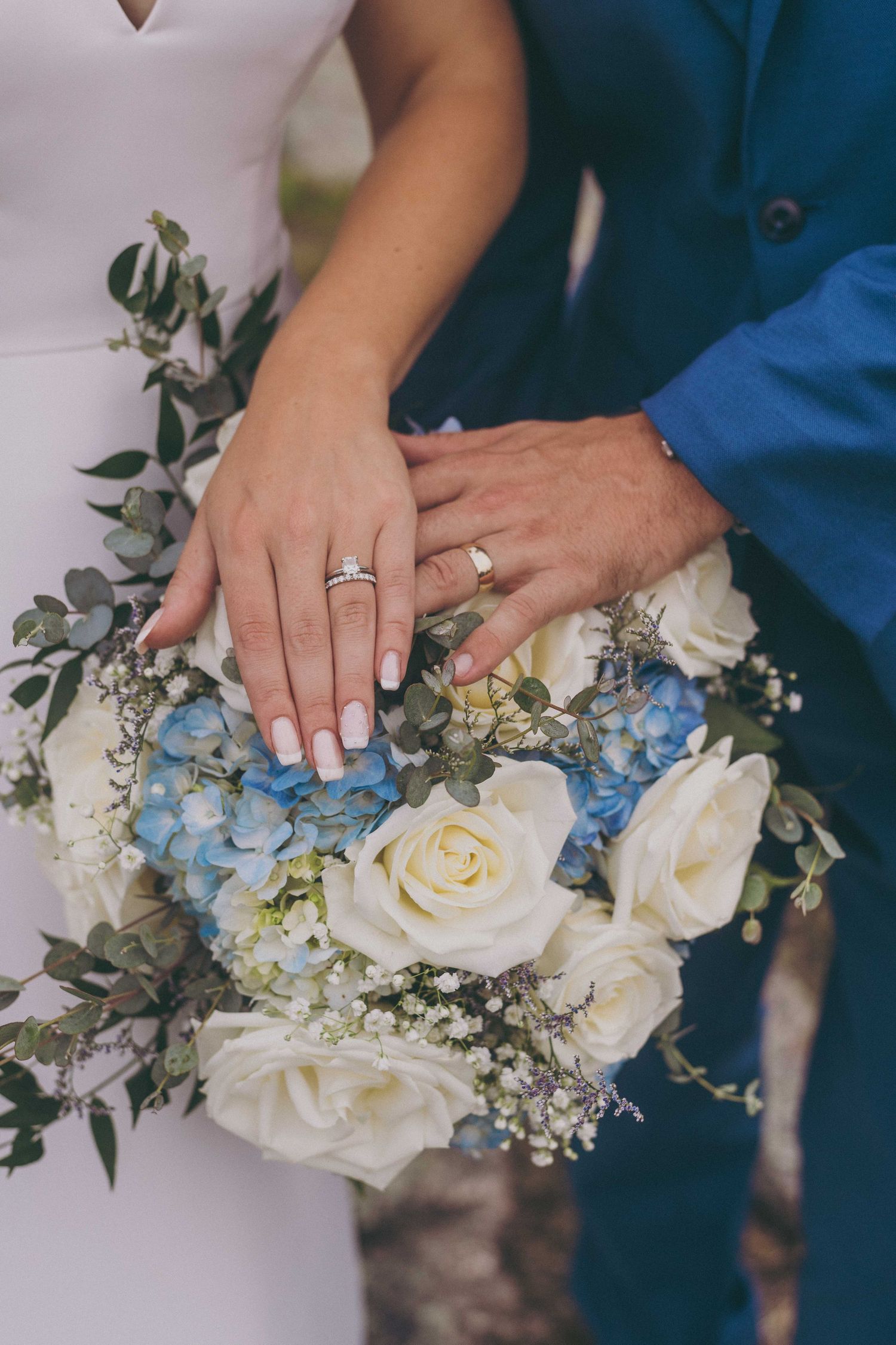 A wedding bouquet of white roses and blue hydrangeas with couple's hands displaying their rings.