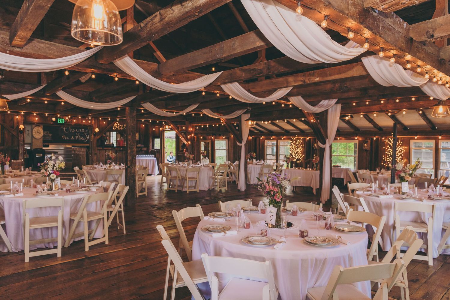 Rustic barn wedding reception venue with white tables, draped fabric, and twinkling lights on wooden beams.