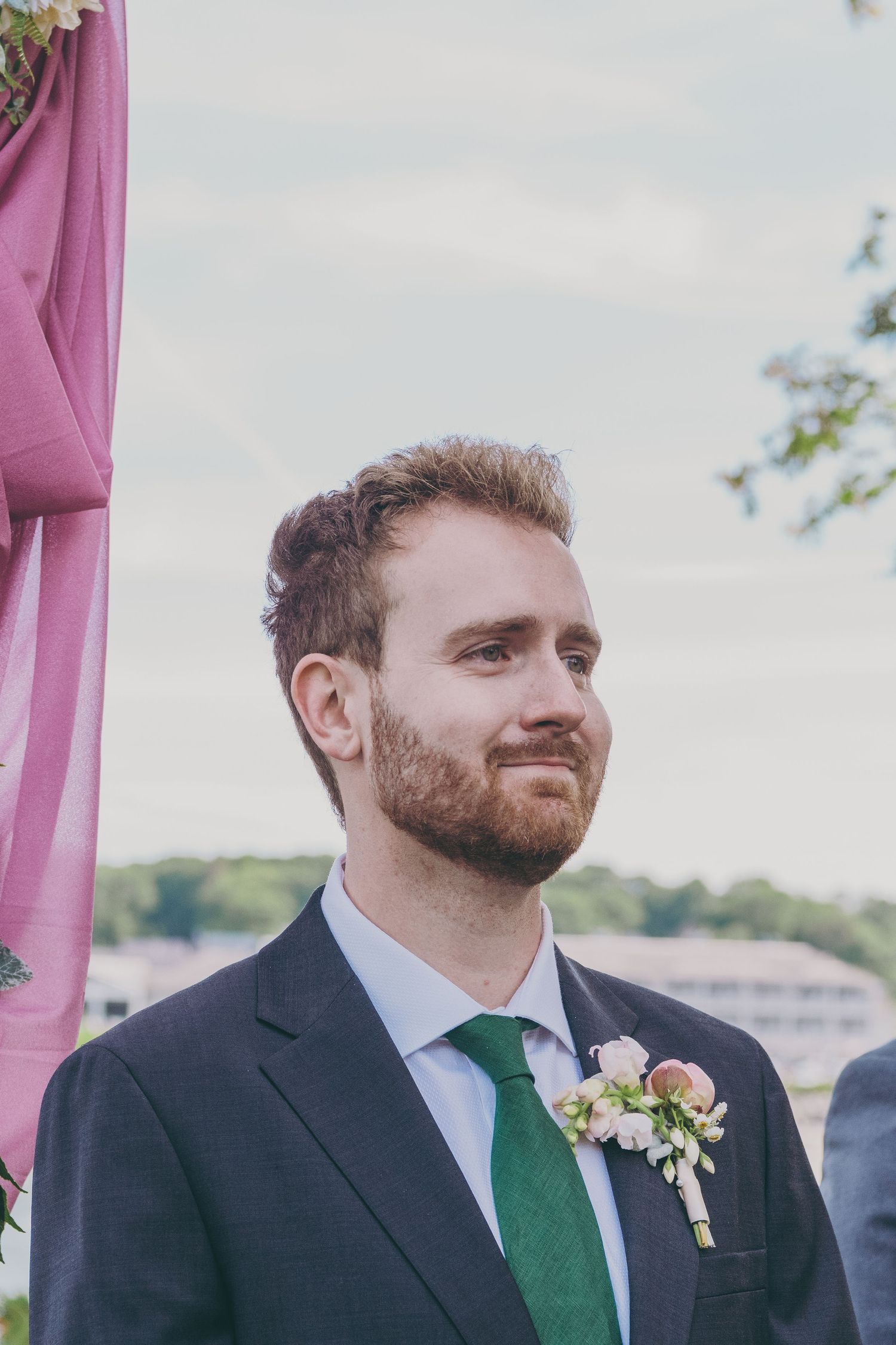 A groom wearing a dark suit and emerald green tie stands at an outdoor wedding ceremony with pink decor visible.