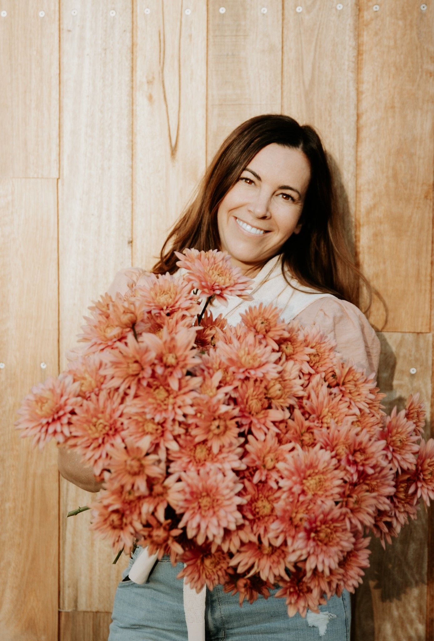 Marielle holding a large bunch of pink chrysanthemums smiling with a backdrop of wooden panels