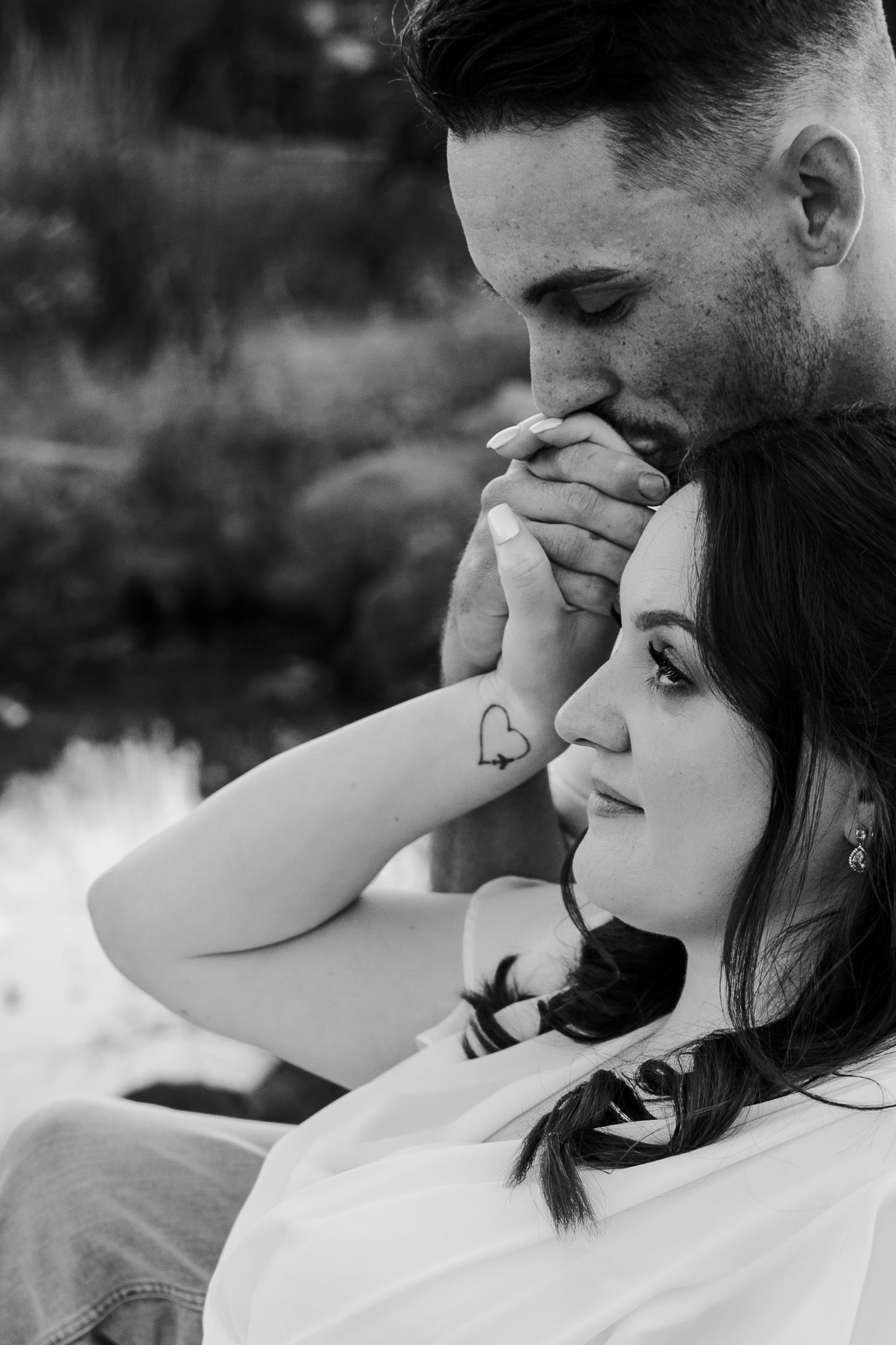 A black and white photo of a newly wed couple, the brides hand is being kissed by the groom as she rests on him.