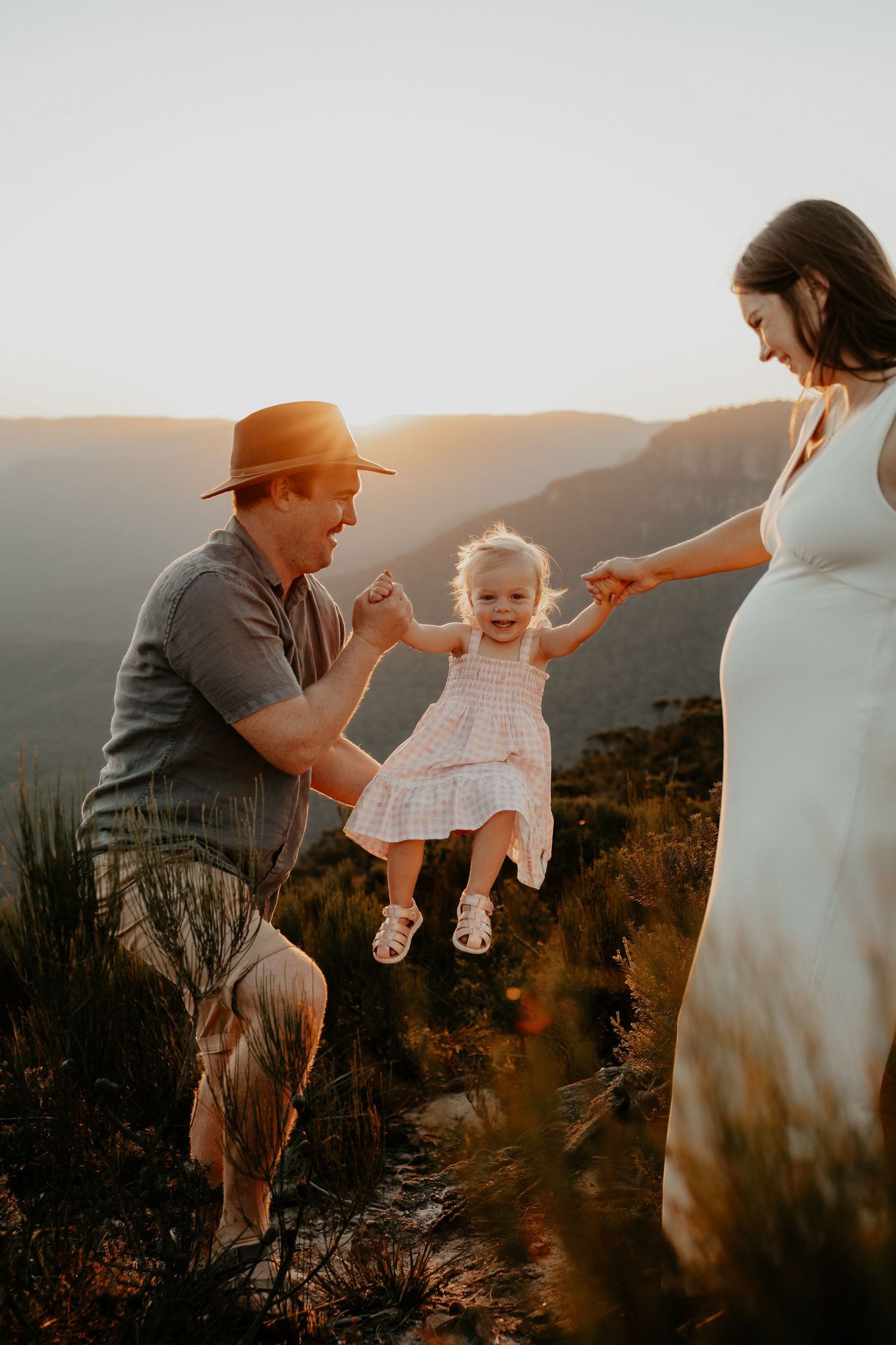 A pregnant mother and her husband both laughing and swinging their young daughter between them at golden hour