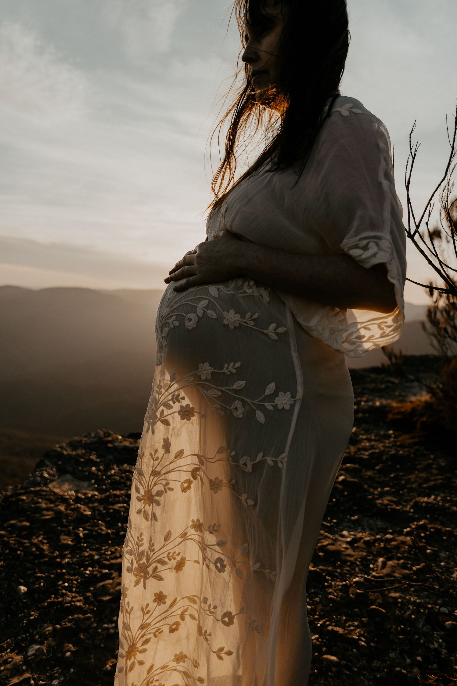 A pregnant mother holding her belly with the sun glowing through her dress emphasizing the silhouette of her pregnant belly.