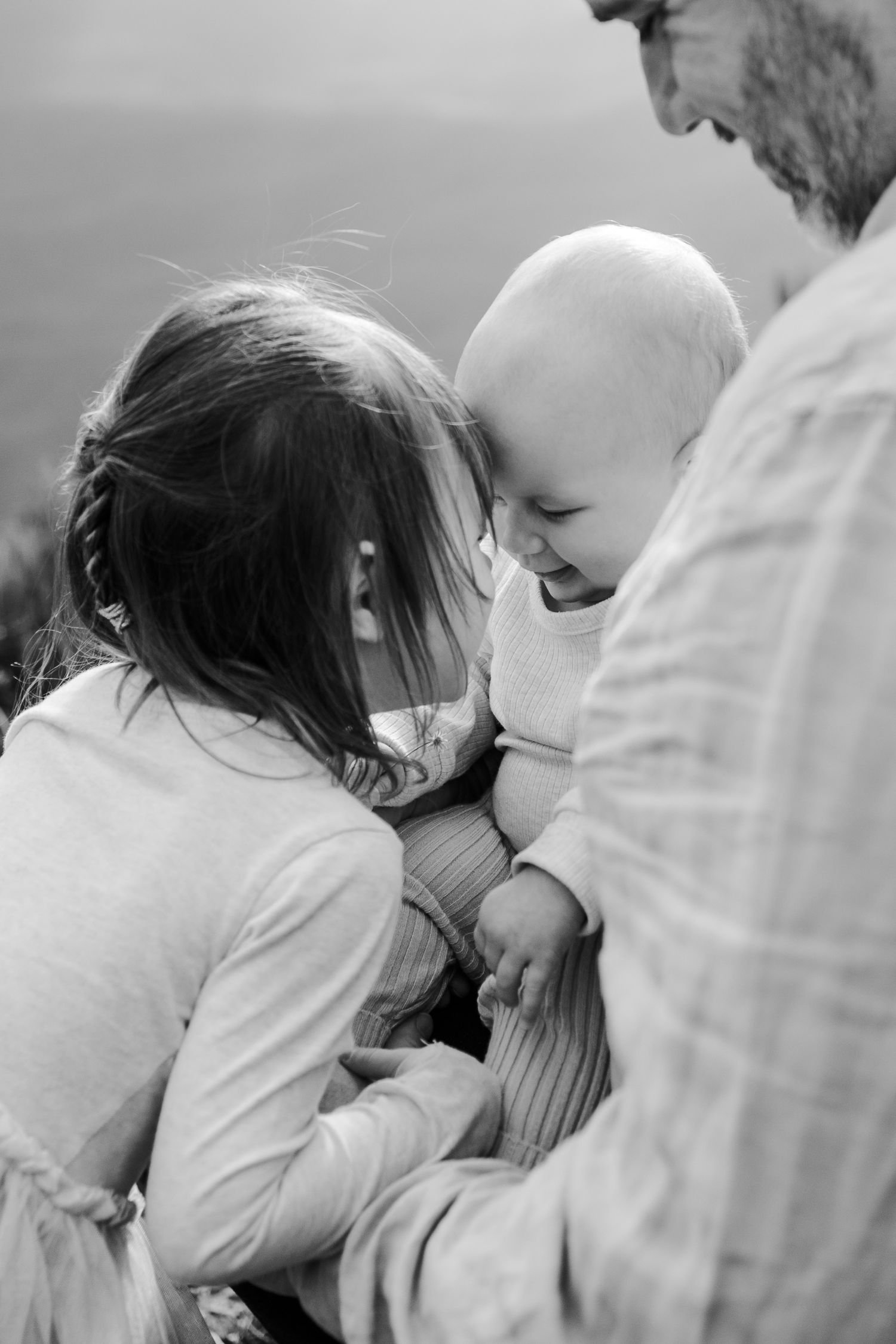 A black and white photo of a baby sitting on her fathers lap with her sister resting her face on her forehead while smiling