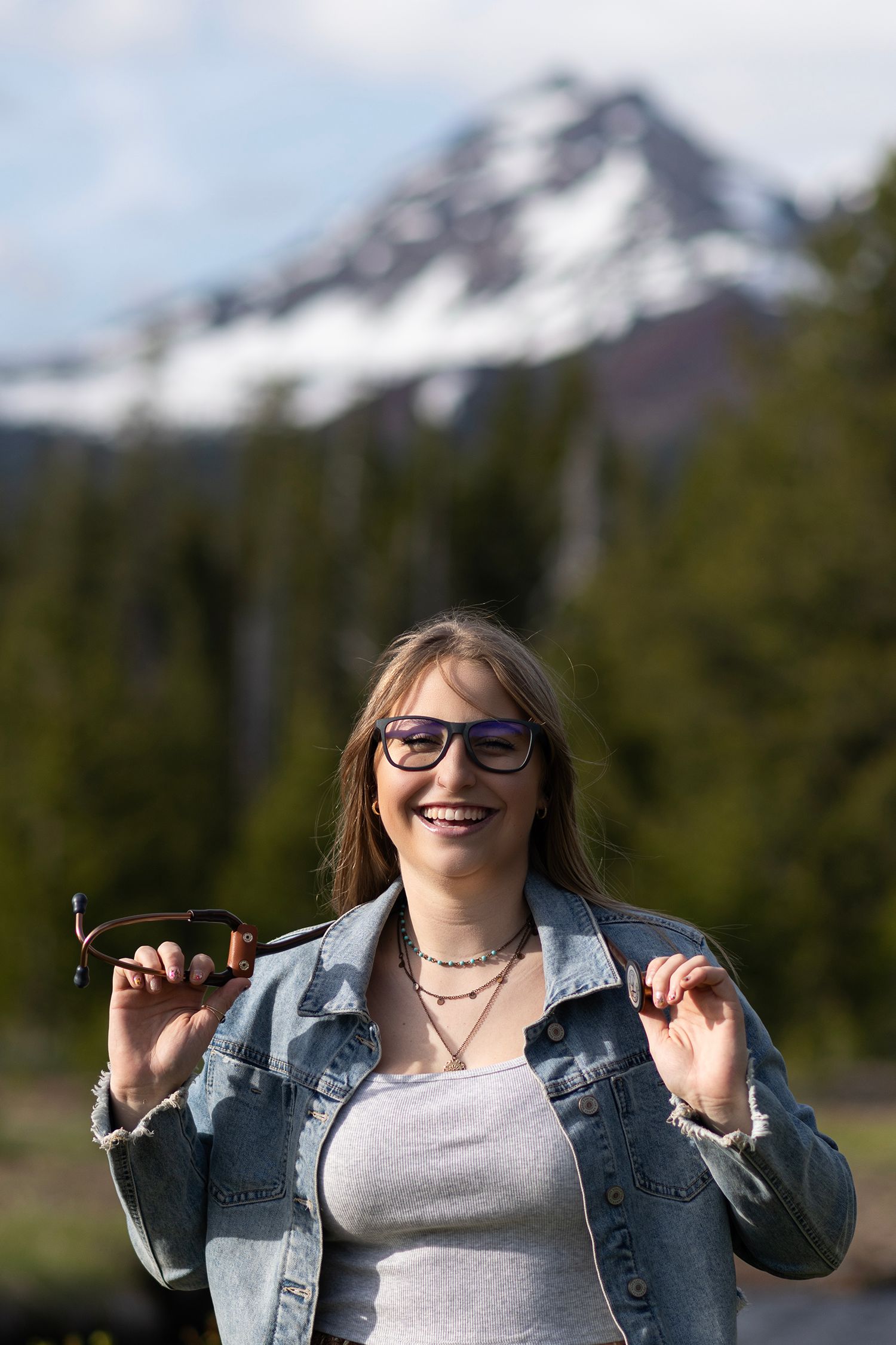 Smiling young woman in denim jacket holding sunglasses with snow-capped mountain and forest backdrop.