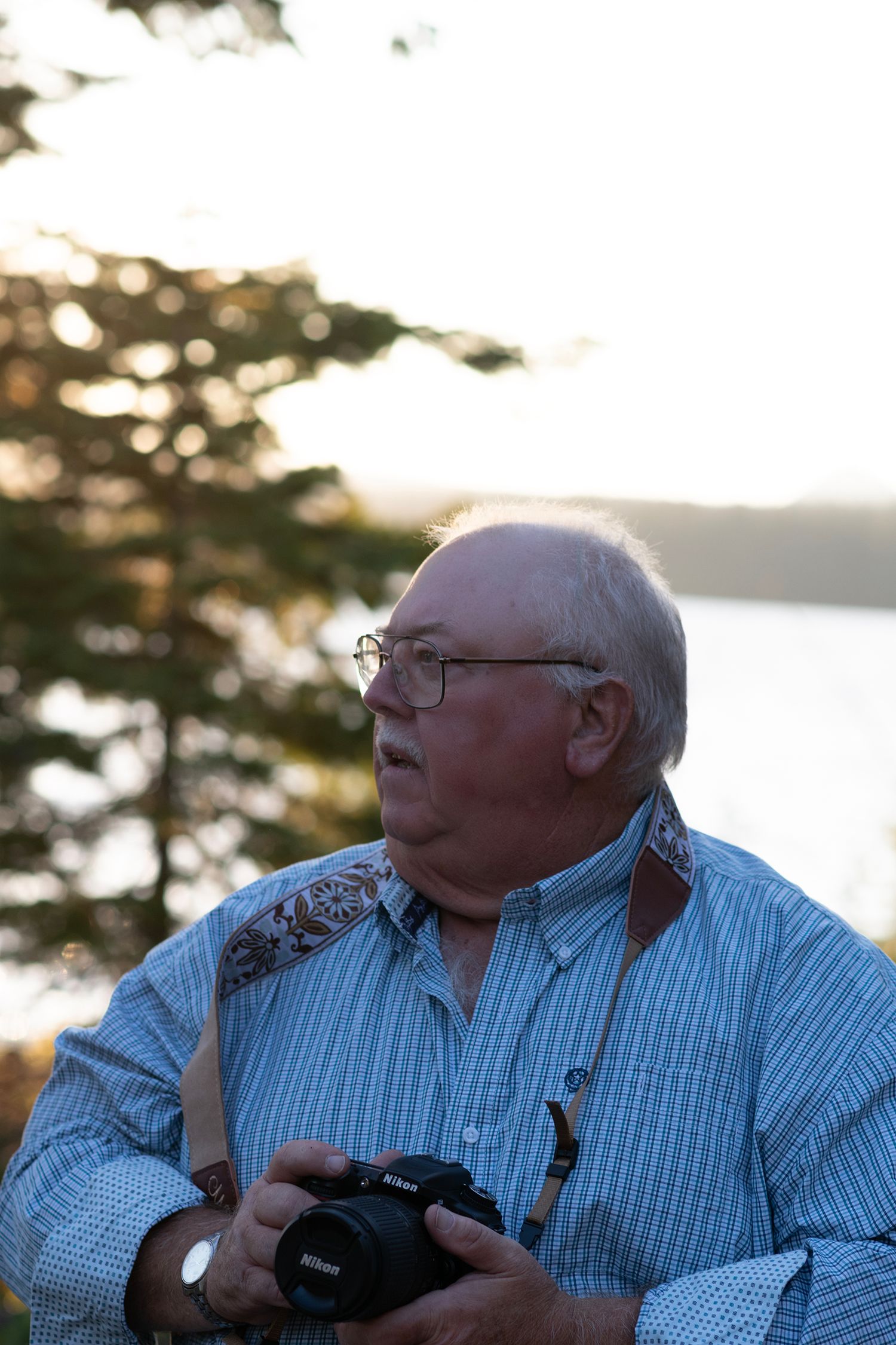 An elderly person wearing a blue checkered shirt holds a camera while enjoying the outdoors at sunset.