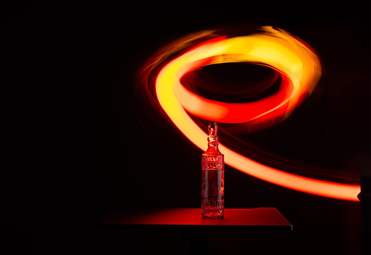 Long exposure light painting creates a glowing orange spiral above a glass bottle against a dark background.