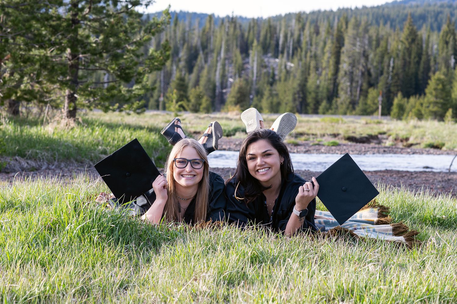 Two graduates lying in a meadow holding diplomas, smiling outdoors with pine trees and a stream in the background.