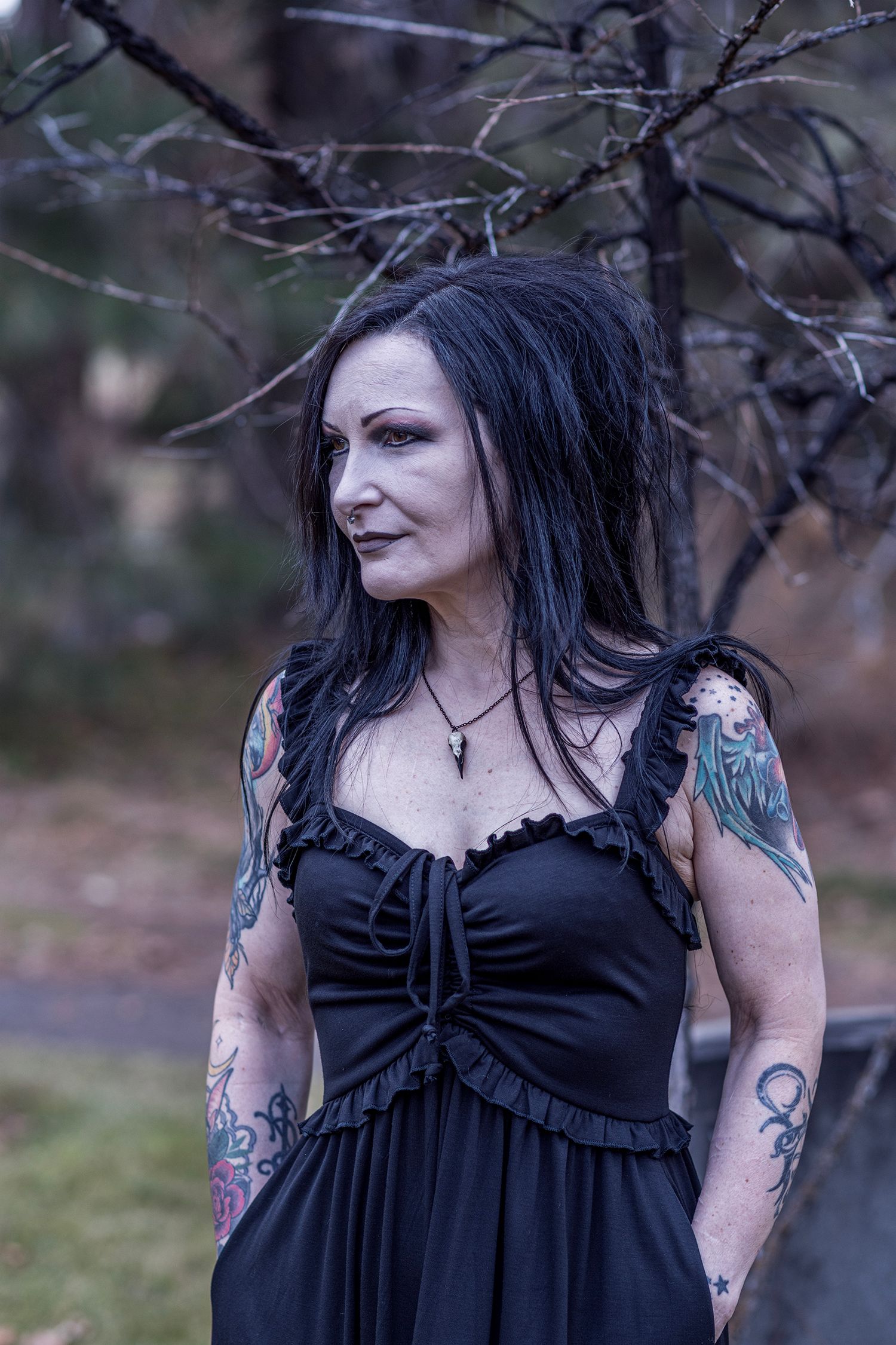 Gothic style portrait in black dress with tattoos standing near bare tree branches in moody outdoor setting.