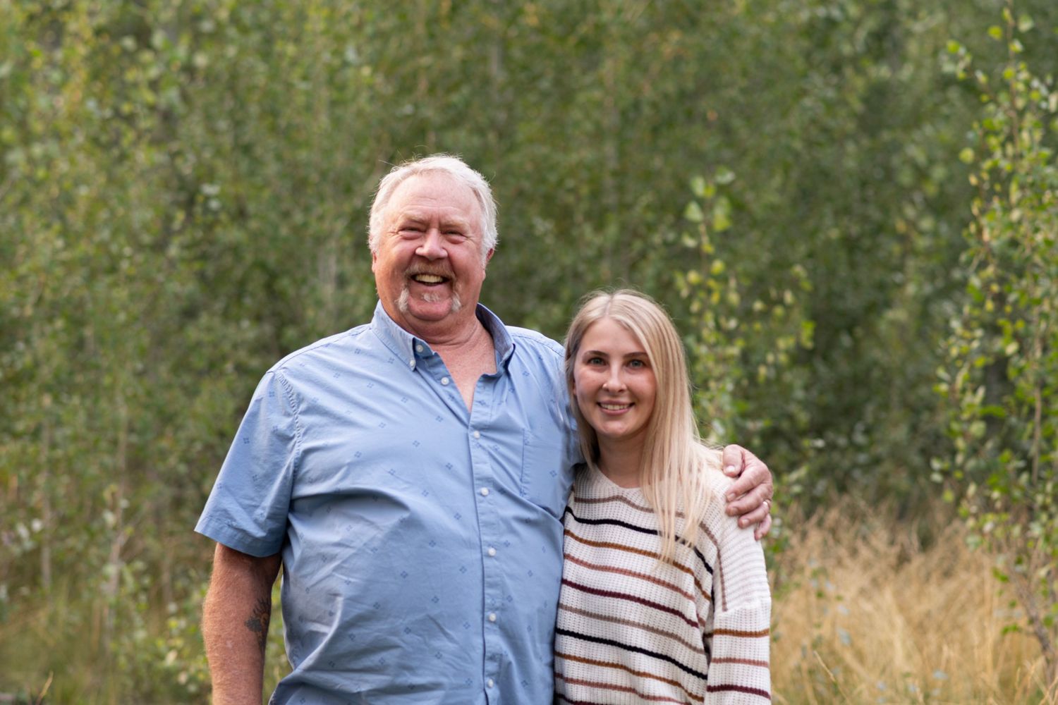 A couple poses together outdoors on a sunny day among tall grass and trees in an extended photo sequence.