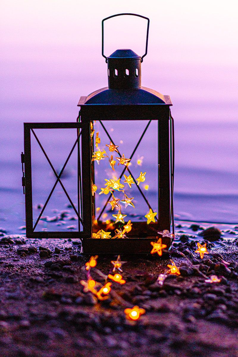 Illuminated lantern with glowing star lights on a beach at dusk creates a magical purple twilight scene.