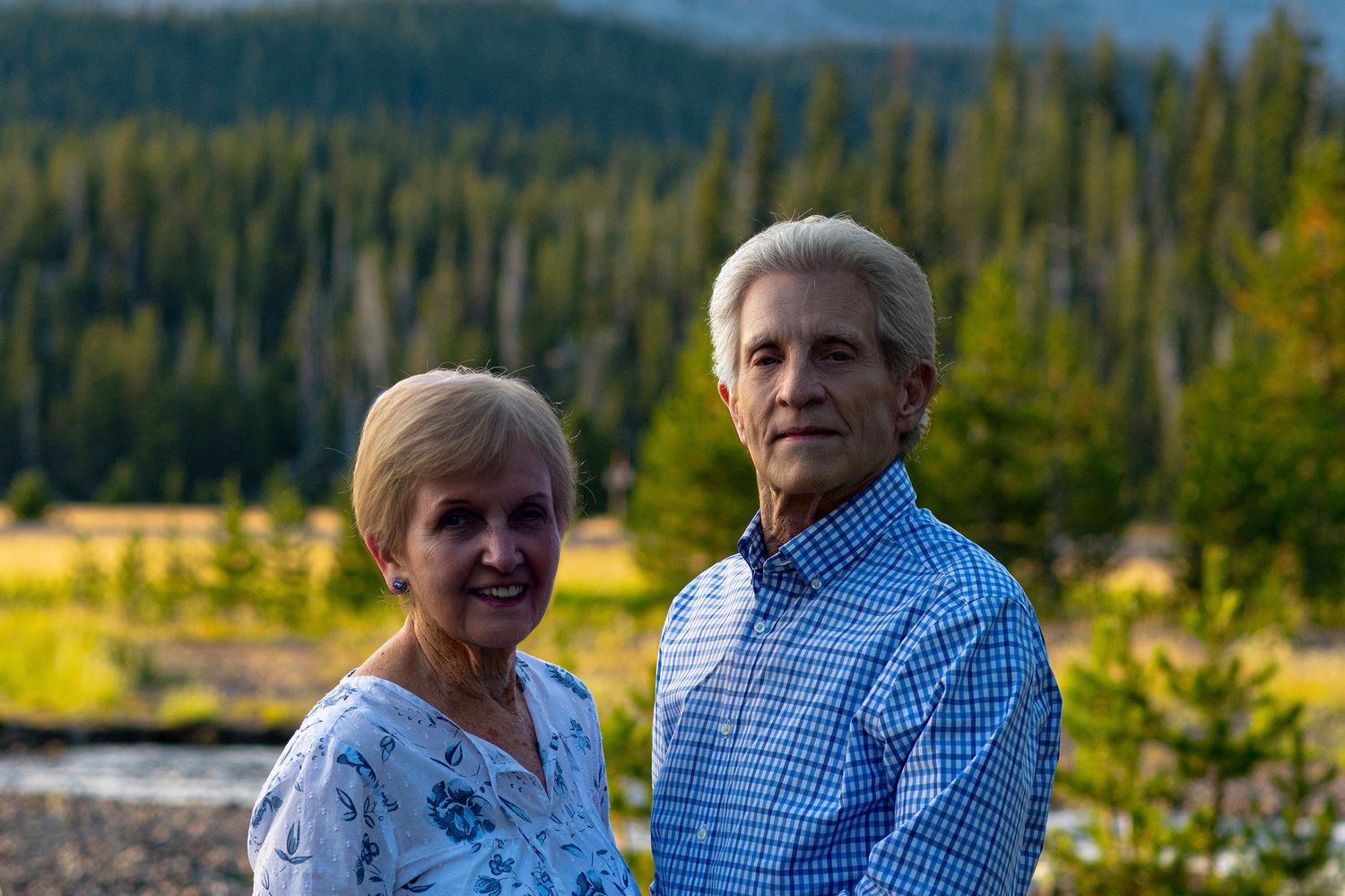 Couple poses together outdoors against a scenic backdrop of evergreen forest and mountains during golden hour.
