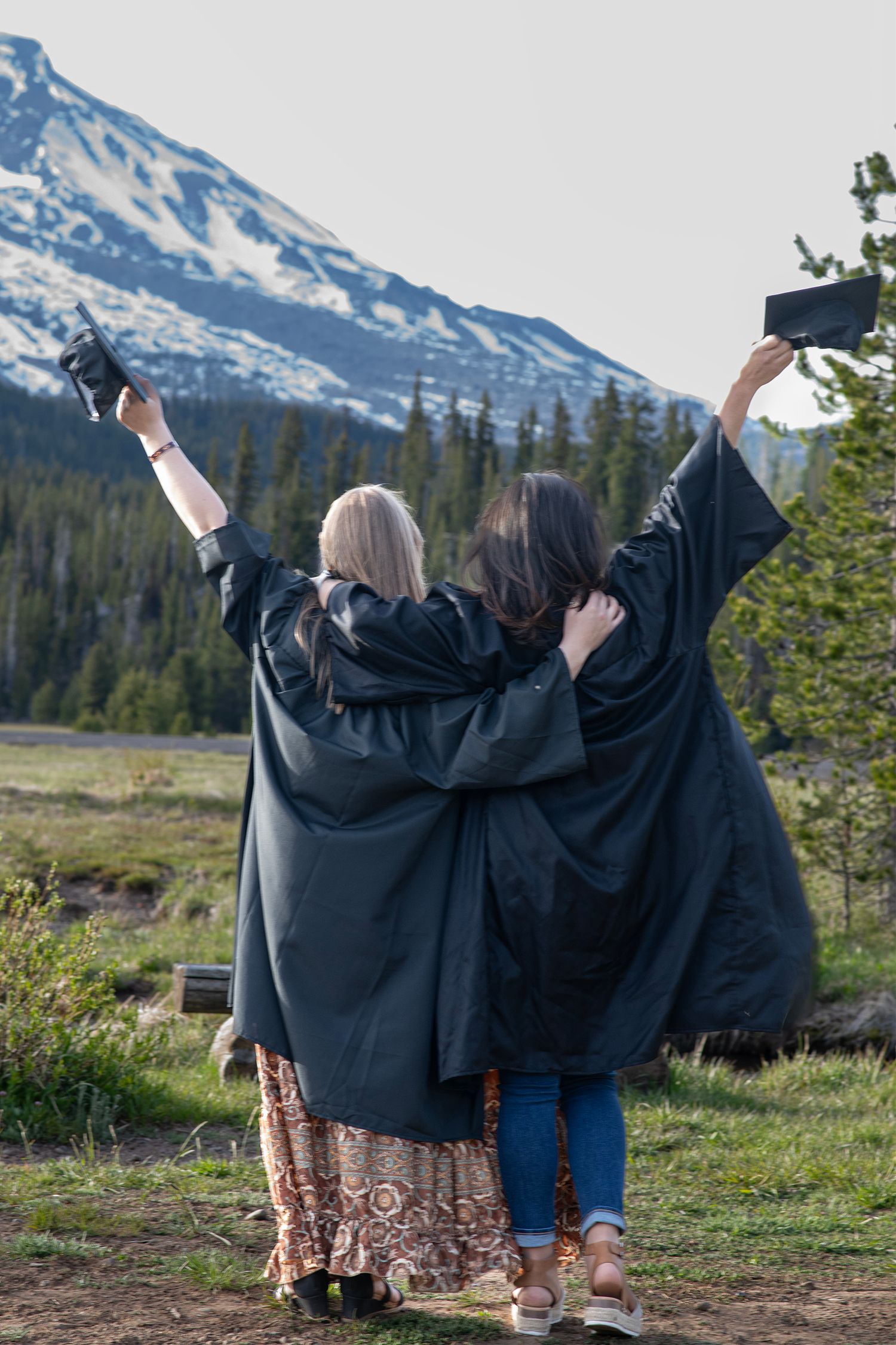 Two graduates in black robes celebrating with arms raised facing snow-capped mountain in scenic outdoor setting.