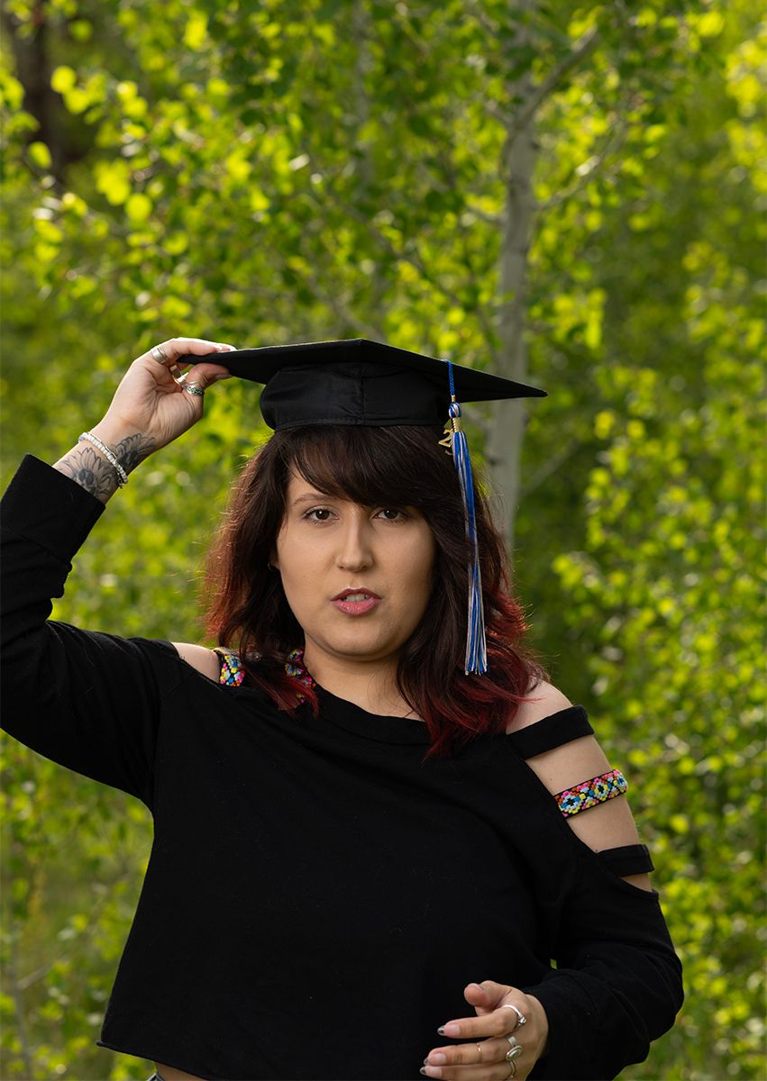Graduate in black dress and cap posing against green foliage background.