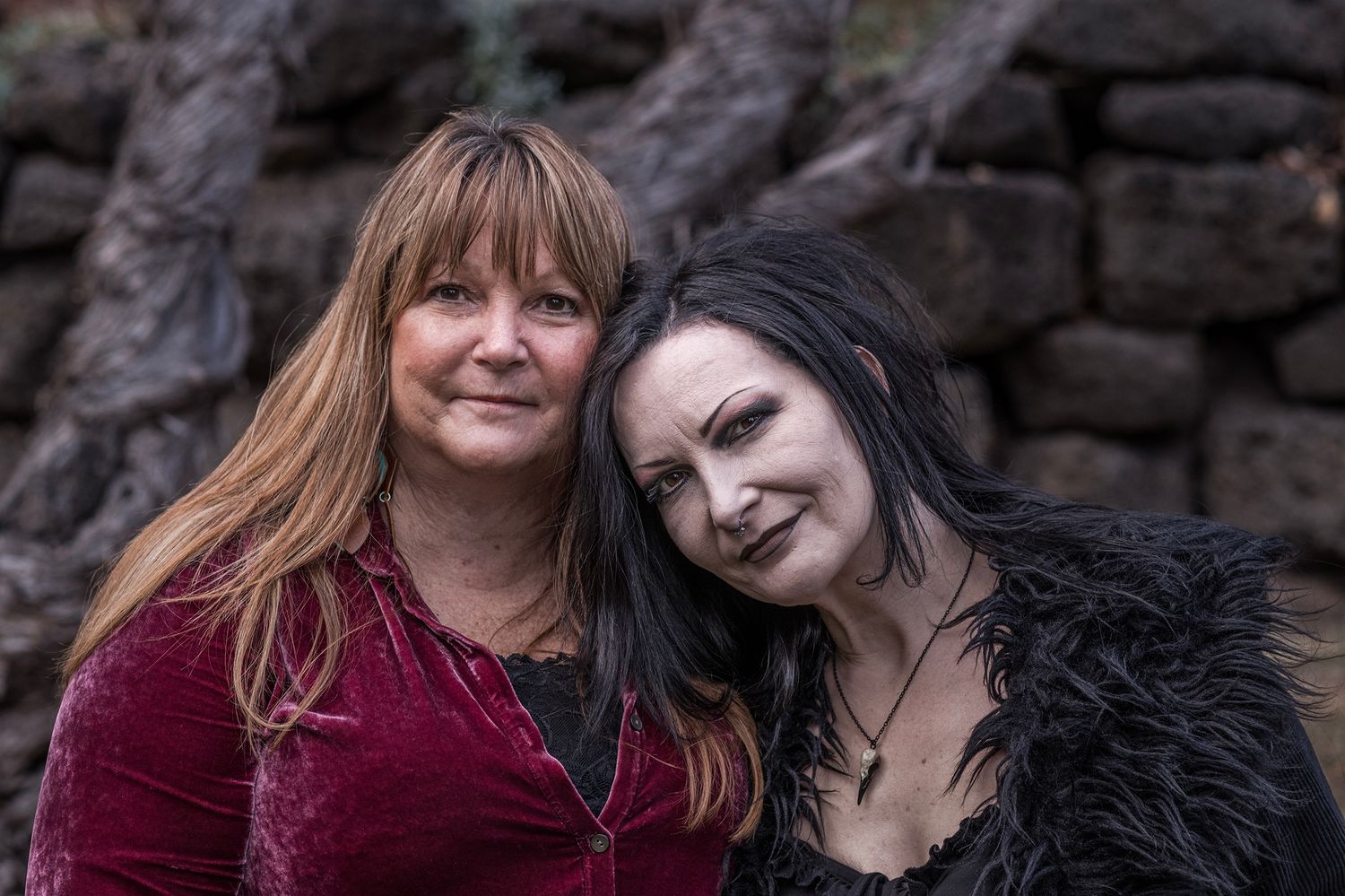 Two people with contrasting hair colors smile together in front of a stone wall.