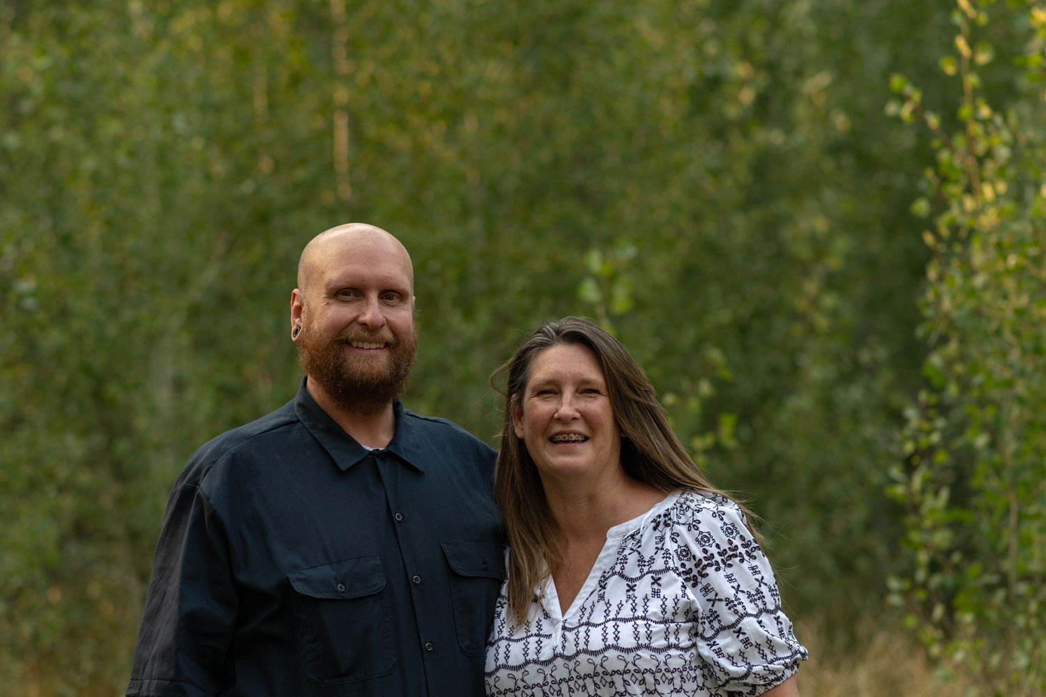 A couple poses together outdoors against a green forest background on an autumn evening.