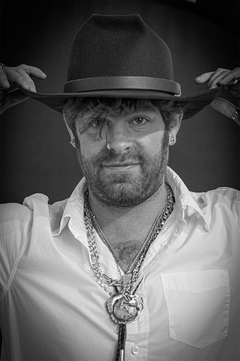 Black and white portrait of a person adjusting their wide-brimmed hat with western-style accessories.