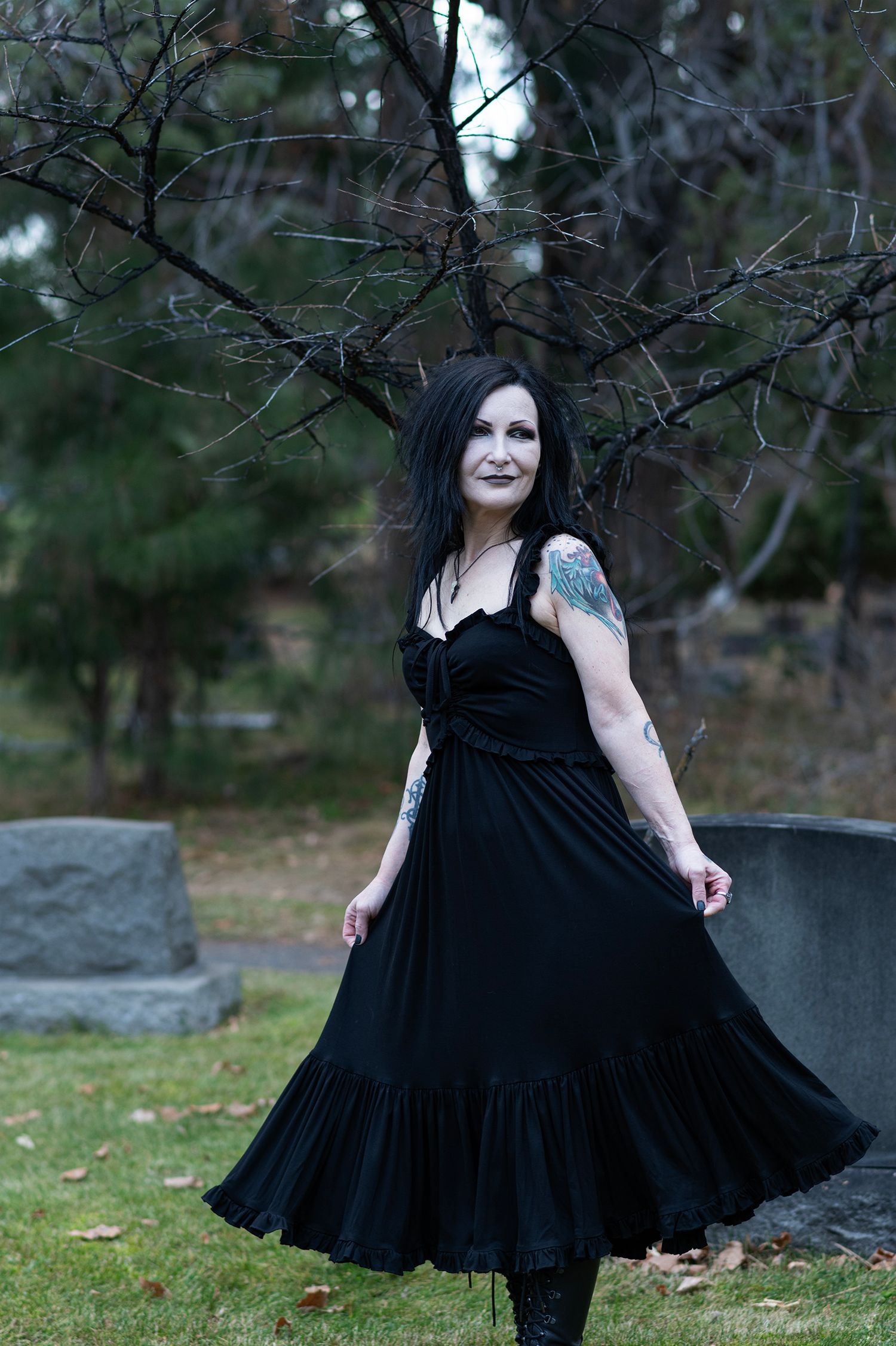 A person wearing a flowing black gothic dress poses among bare tree branches in a cemetery setting.