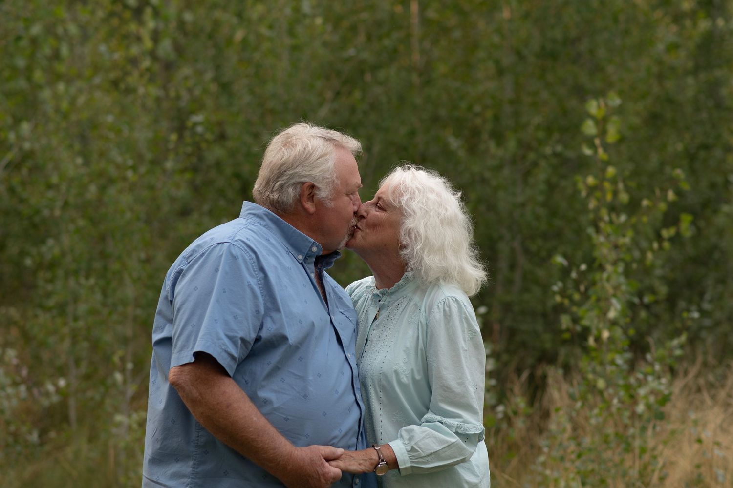 A loving elderly couple share tender kisses while walking in a lush green natural setting.