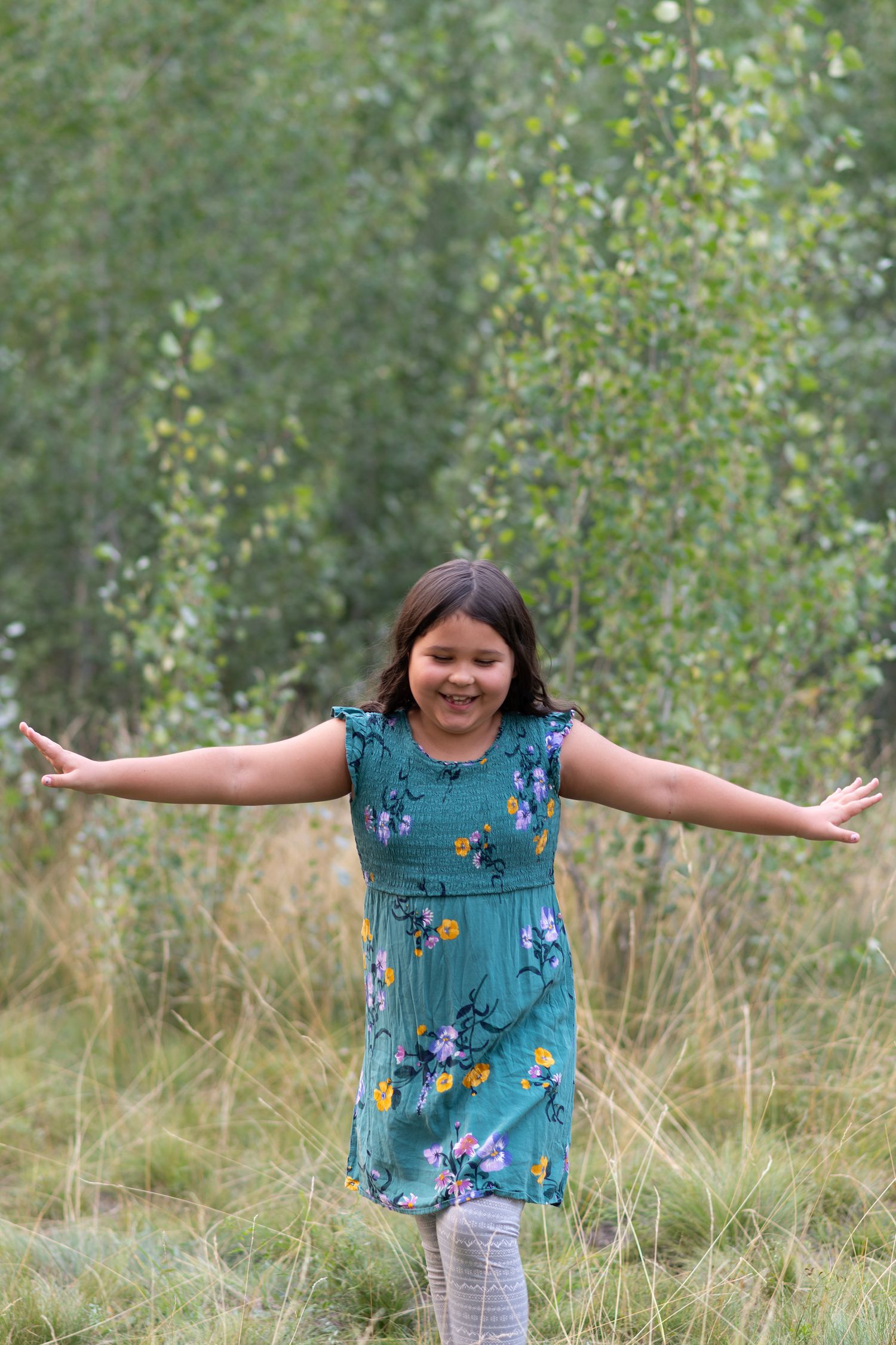 A child wearing a blue floral dress poses joyfully with arms outstretched in a grassy outdoor setting.
