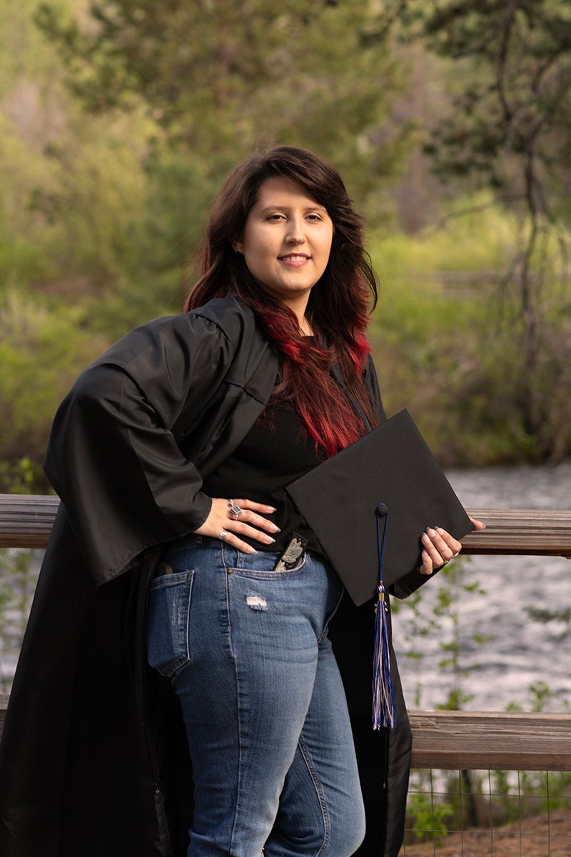 Graduate in black academic regalia poses with diploma on wooden bridge outdoors.