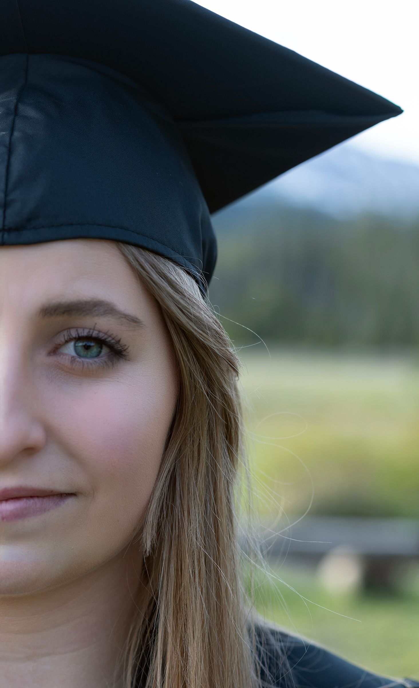 Young woman in black graduation cap smiling outdoors with green landscape and mountains in background.