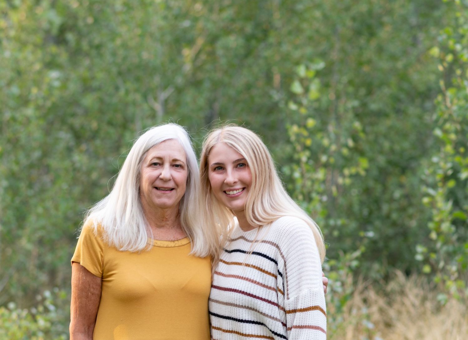 Two people wearing yellow and striped shirts embrace and laugh while standing outdoors in a natural setting.