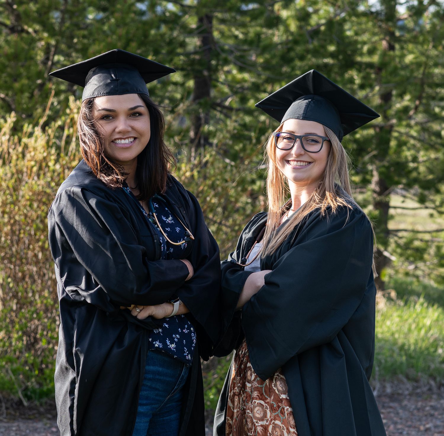 Two smiling women in black graduation caps and gowns pose with arms crossed outdoors near green trees.