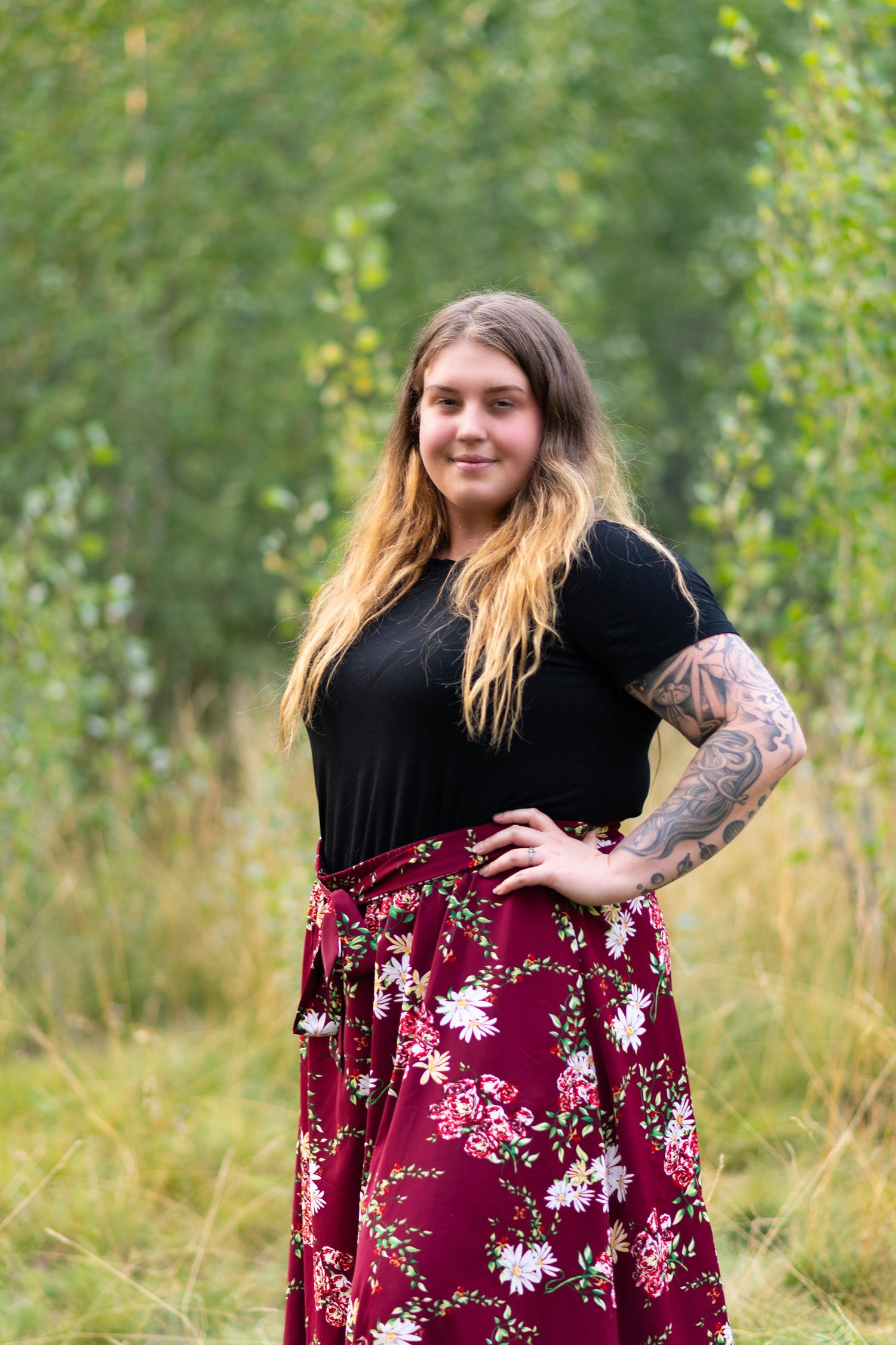 A woman in a black top and burgundy floral maxi skirt poses outdoors against a natural wooded background.