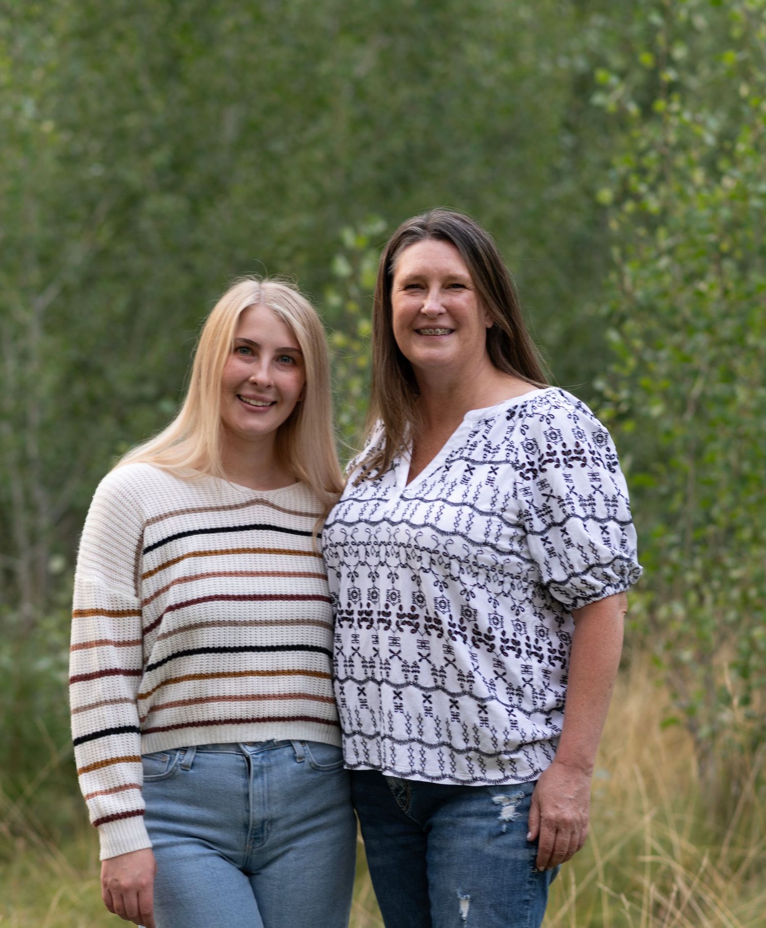 Two women in casual outfits stand together outdoors with green foliage in the background.