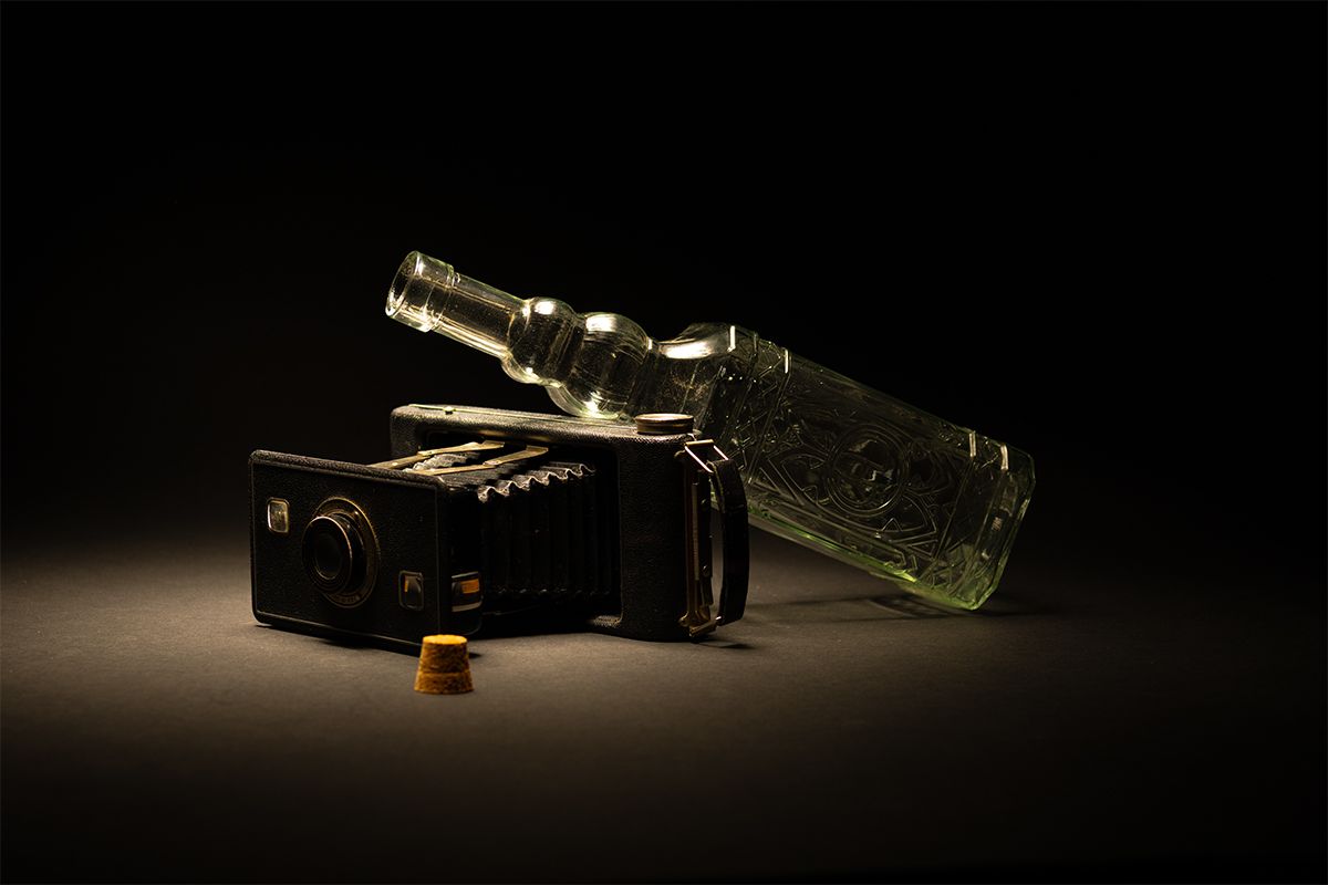 A vintage camera and glass bottle arranged artistically on dark background with dramatic lighting.