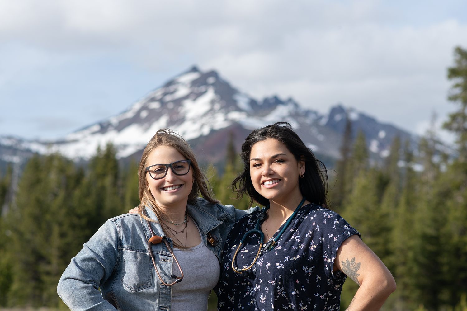 Two women smiling outdoors with a snow-capped mountain and pine trees in the background.