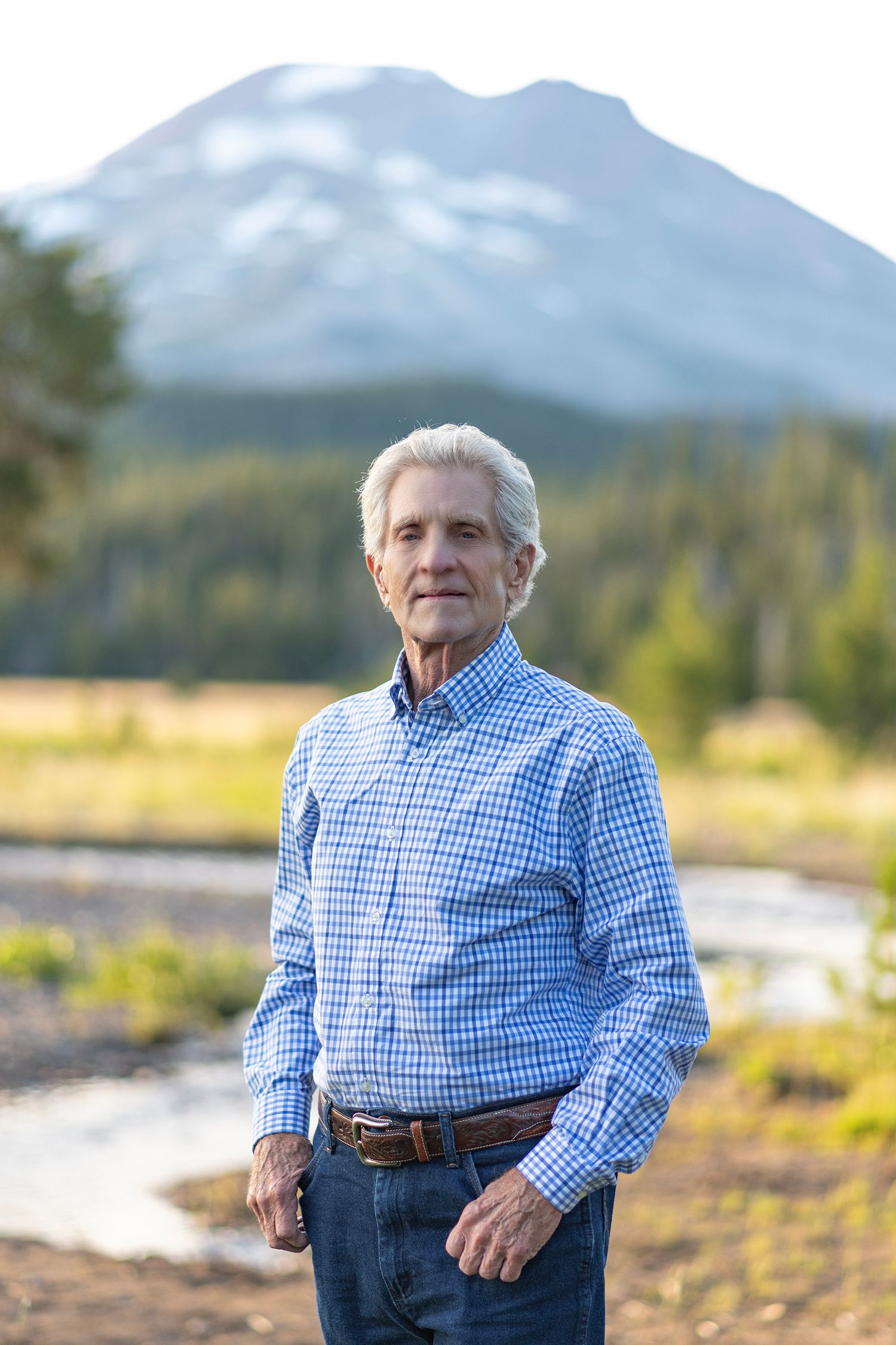 A person in a blue checkered shirt and jeans stands outdoors with a majestic mountain peak rising in the background.