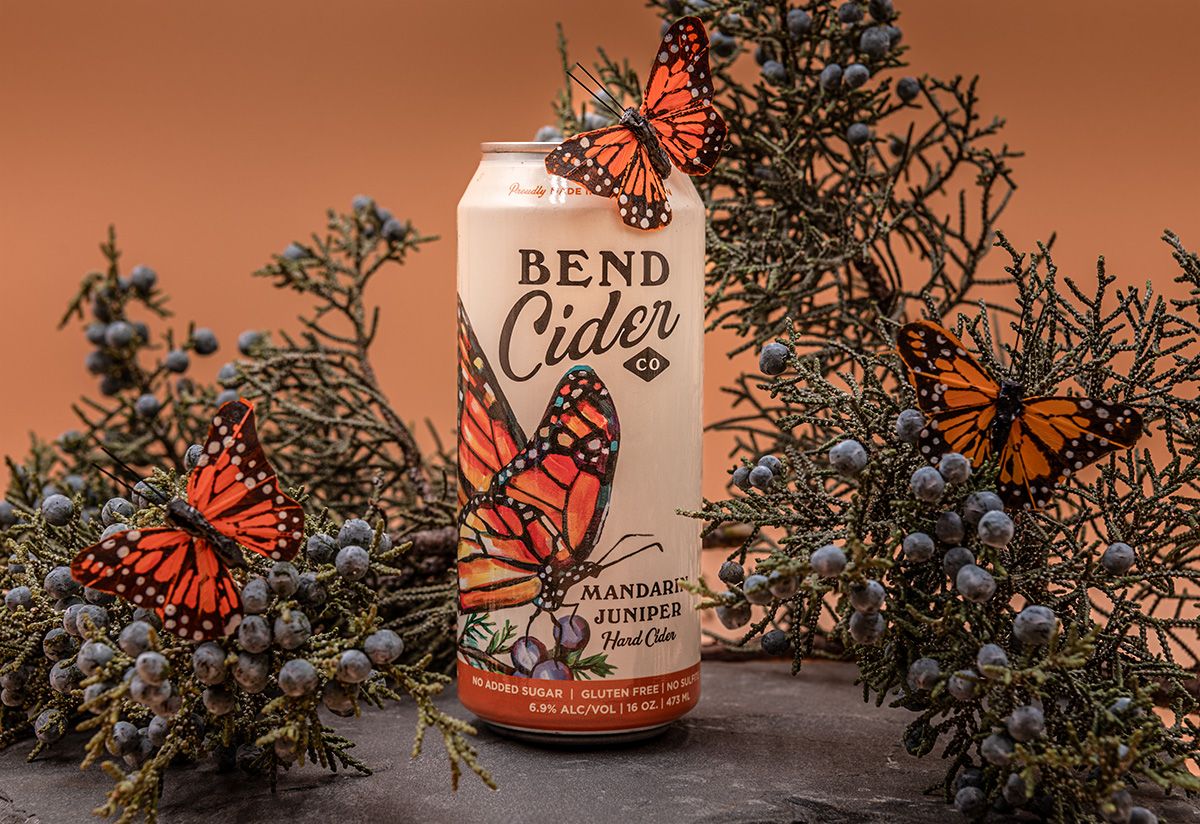 Bend Cider Co. Mandarin Juniper hard cider can surrounded by monarch butterflies and juniper branches on warm orange background.