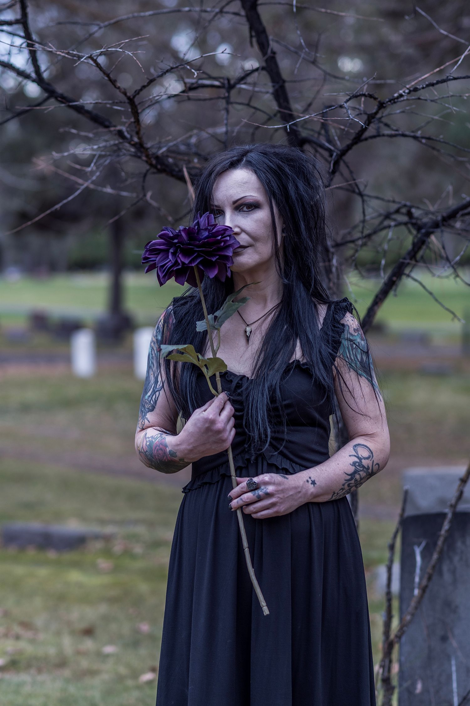 A person in a black gothic dress holds a dark purple flower in a moody outdoor setting with bare trees.