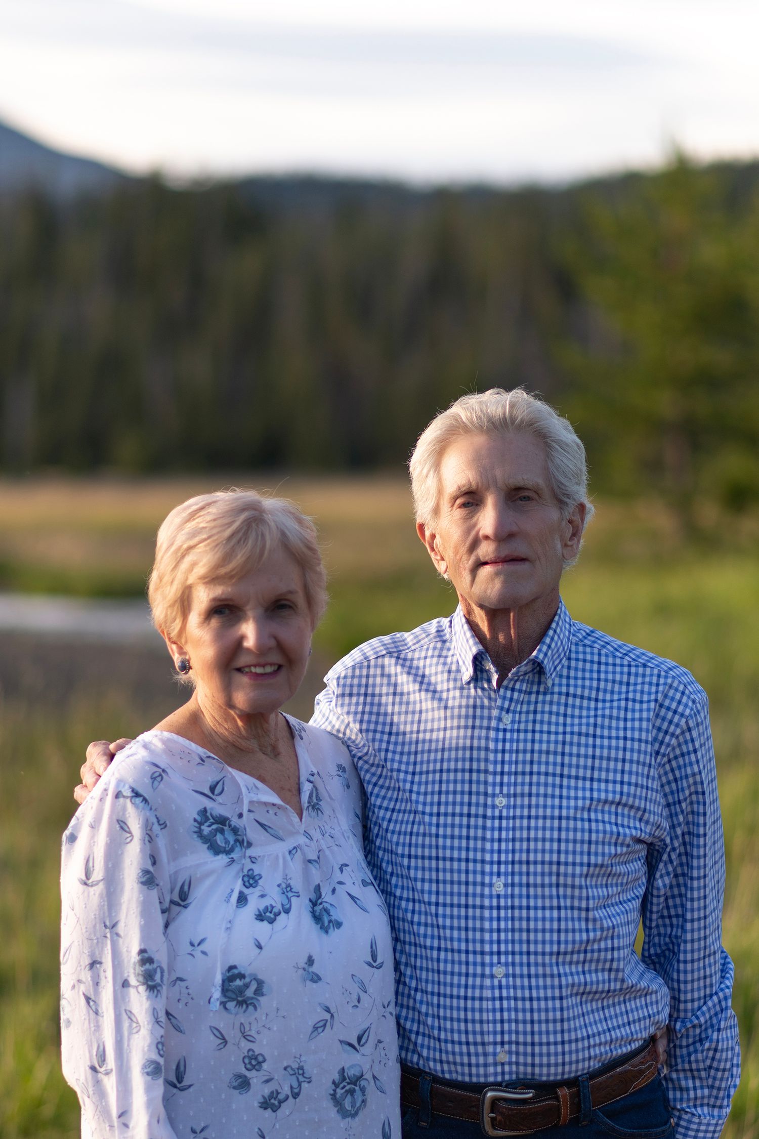 Happy elderly couple embraces while standing outdoors in a grassy field surrounded by mountains and trees.