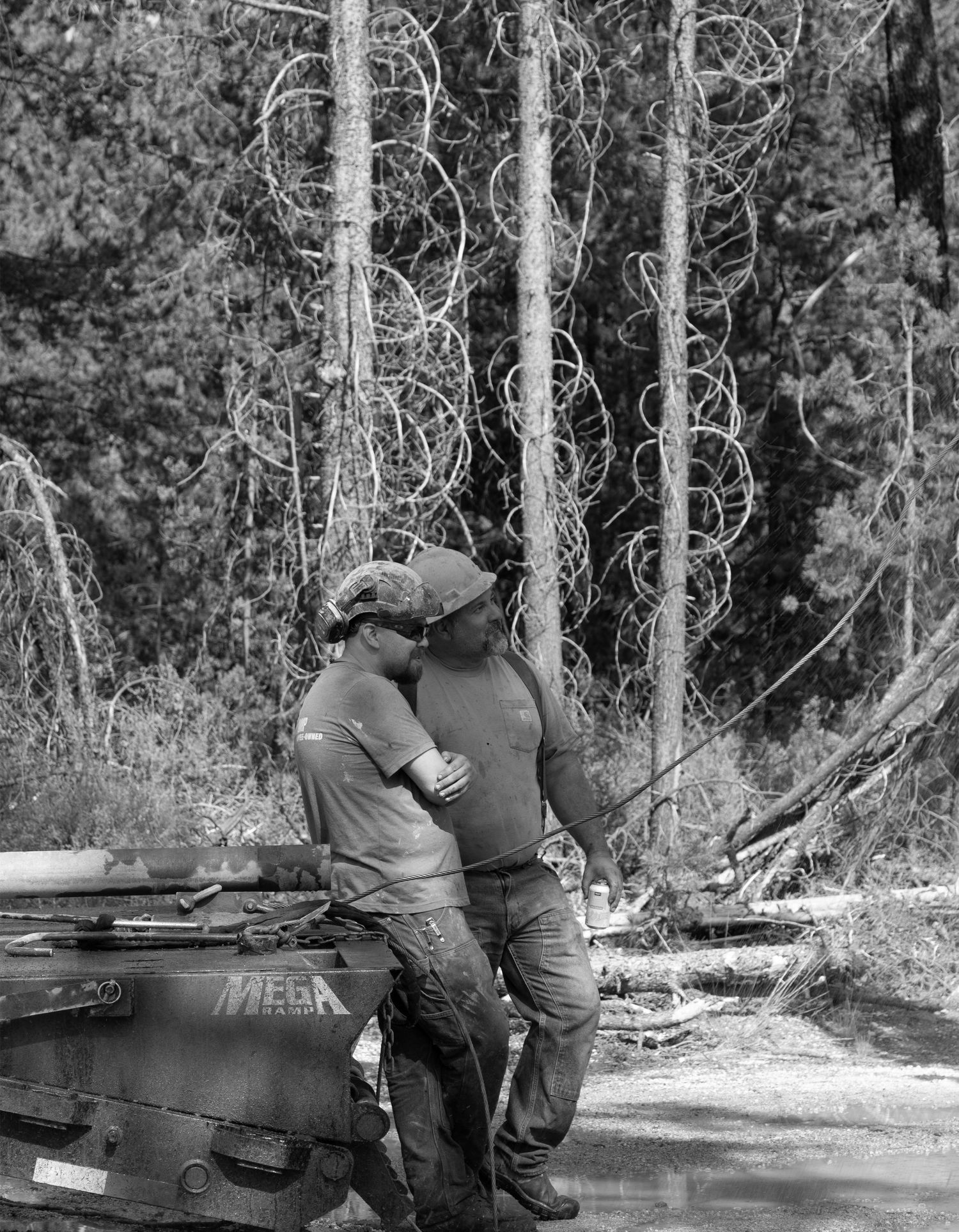 Two figures in casual attire lean against a boat in black and white forest setting.
