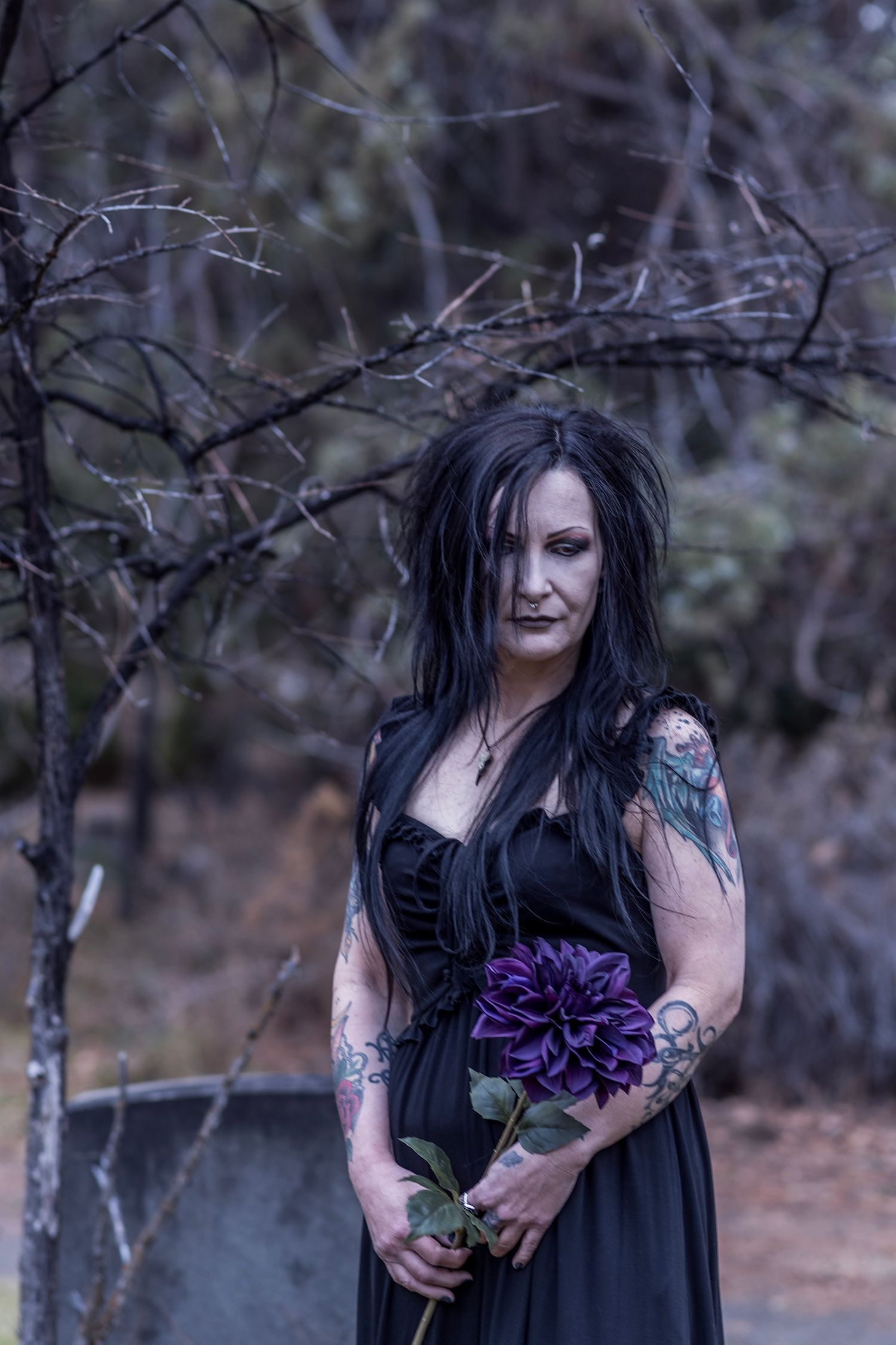 A figure in a black dress holds purple flowers while standing in a moody forest setting.