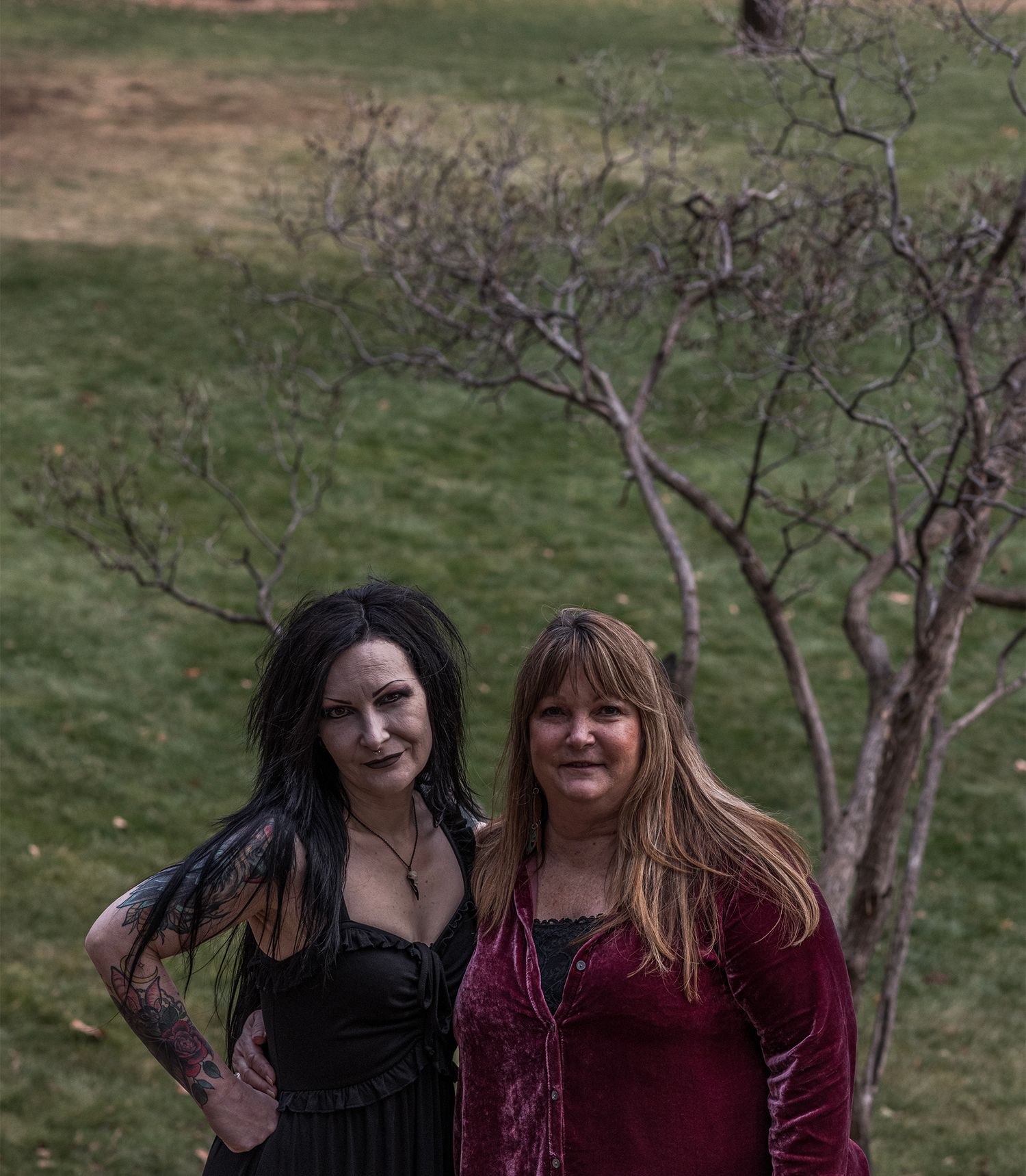 Two people wearing black and burgundy pose together on a grassy hillside with bare trees in the background.