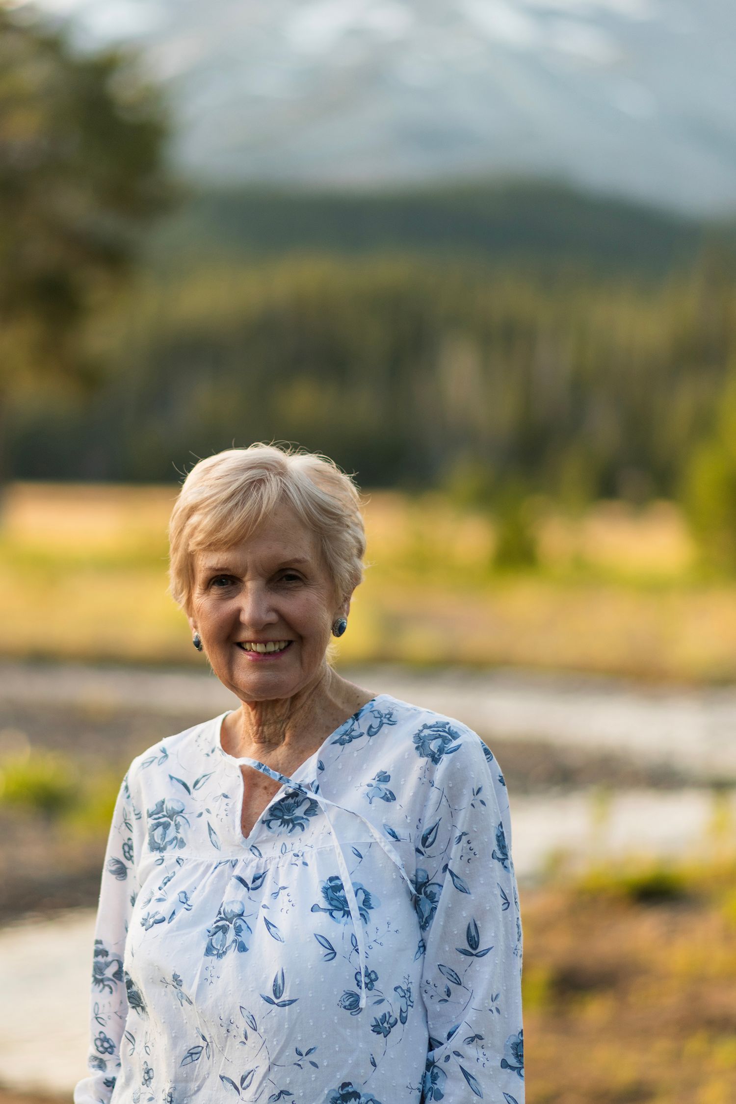 A senior in a white and blue floral blouse stands outdoors against a scenic mountain landscape background.