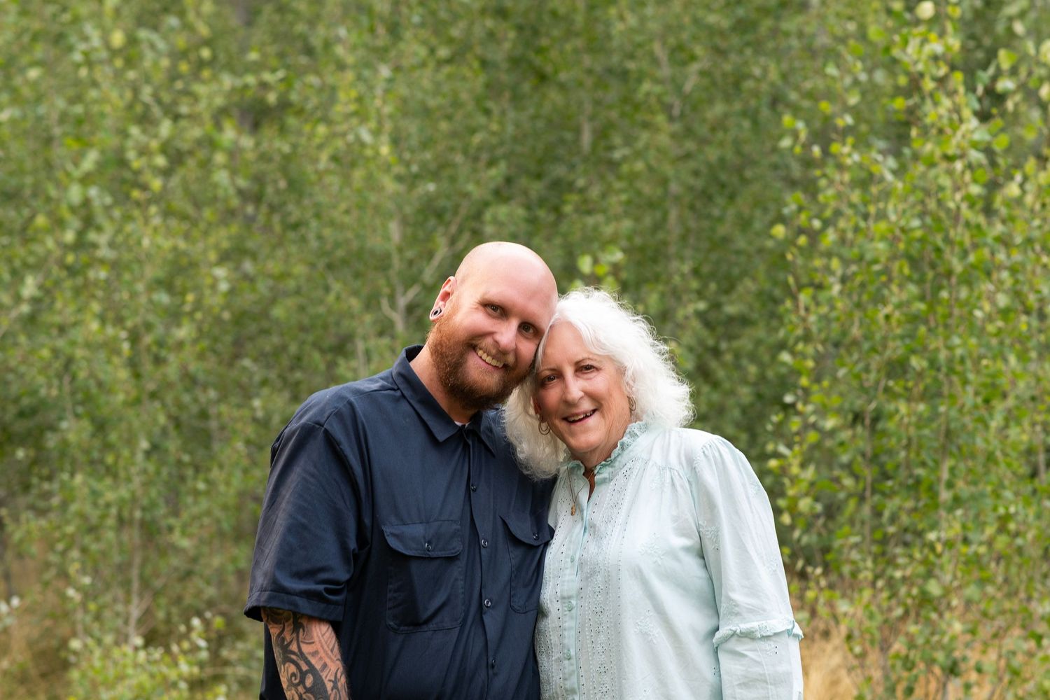A couple poses outdoors smiling together in front of leafy green trees on their wedding day.