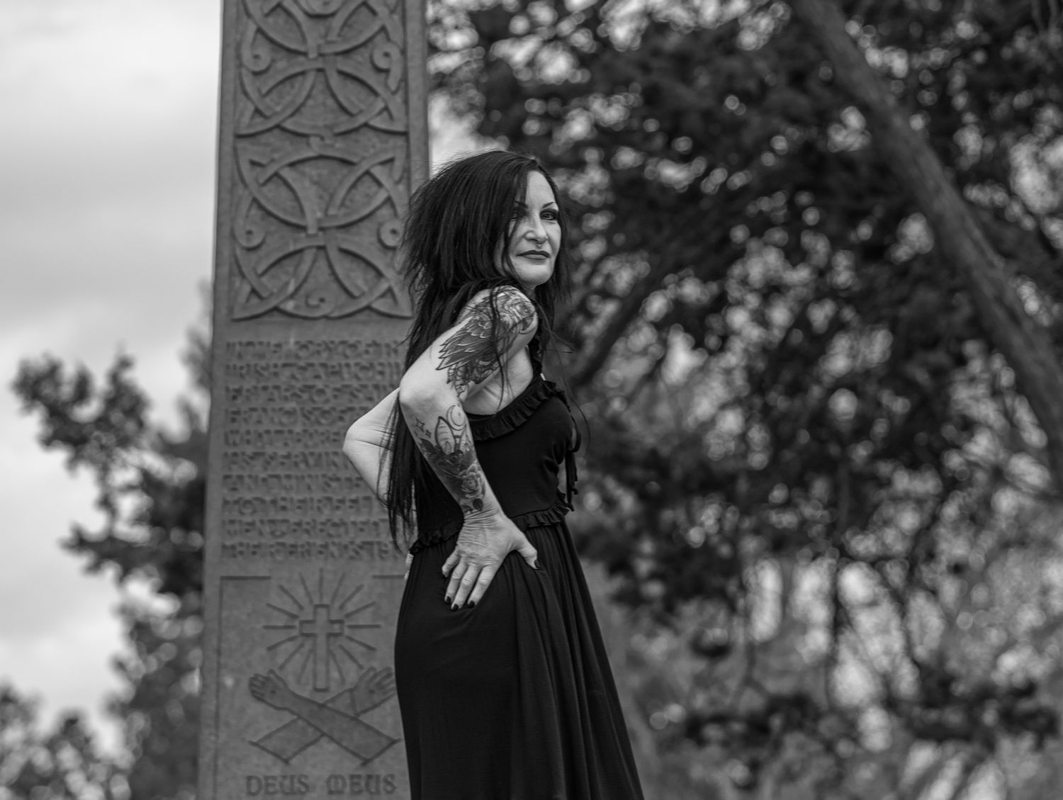 A person in a long black dress poses in front of ornate stone pillars in a black and white photograph.