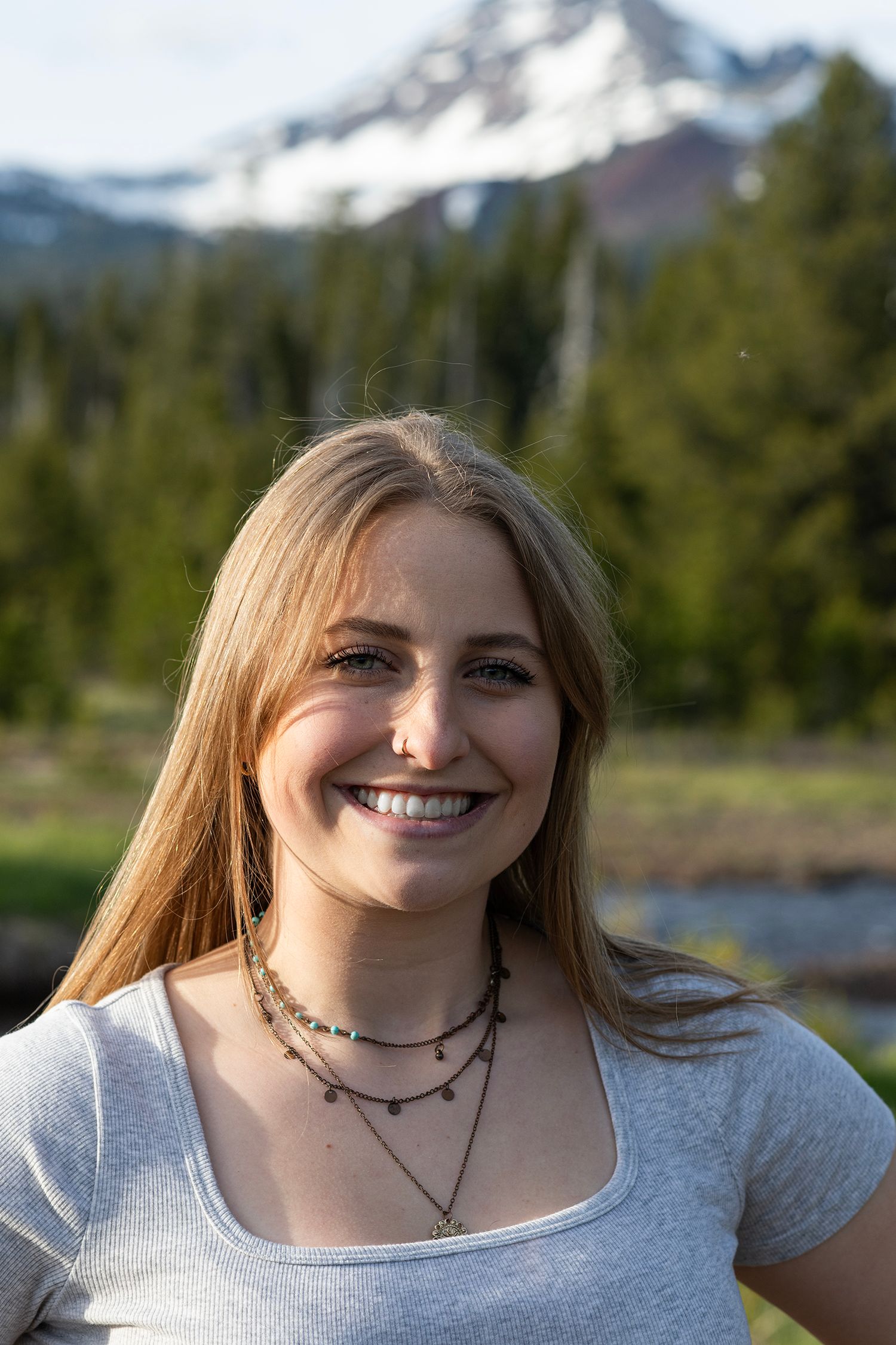 Smiling young woman with long blonde hair wearing layered necklaces outdoors with snowy mountain backdrop.