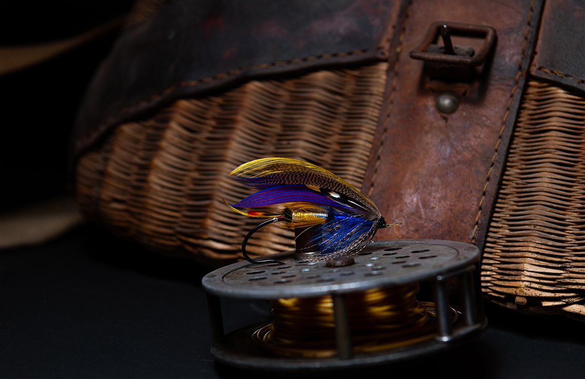 Colorful fly fishing lure resting on a vintage brass reel beside a wicker creel basket.