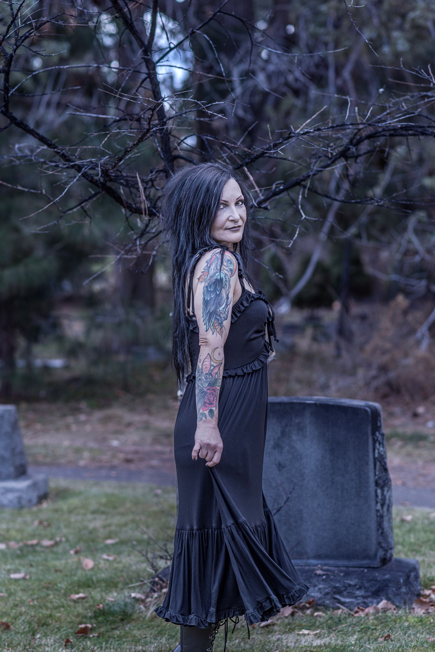 A person in a long black dress stands near a gravestone at dusk, surrounded by bare tree branches in a moody setting.