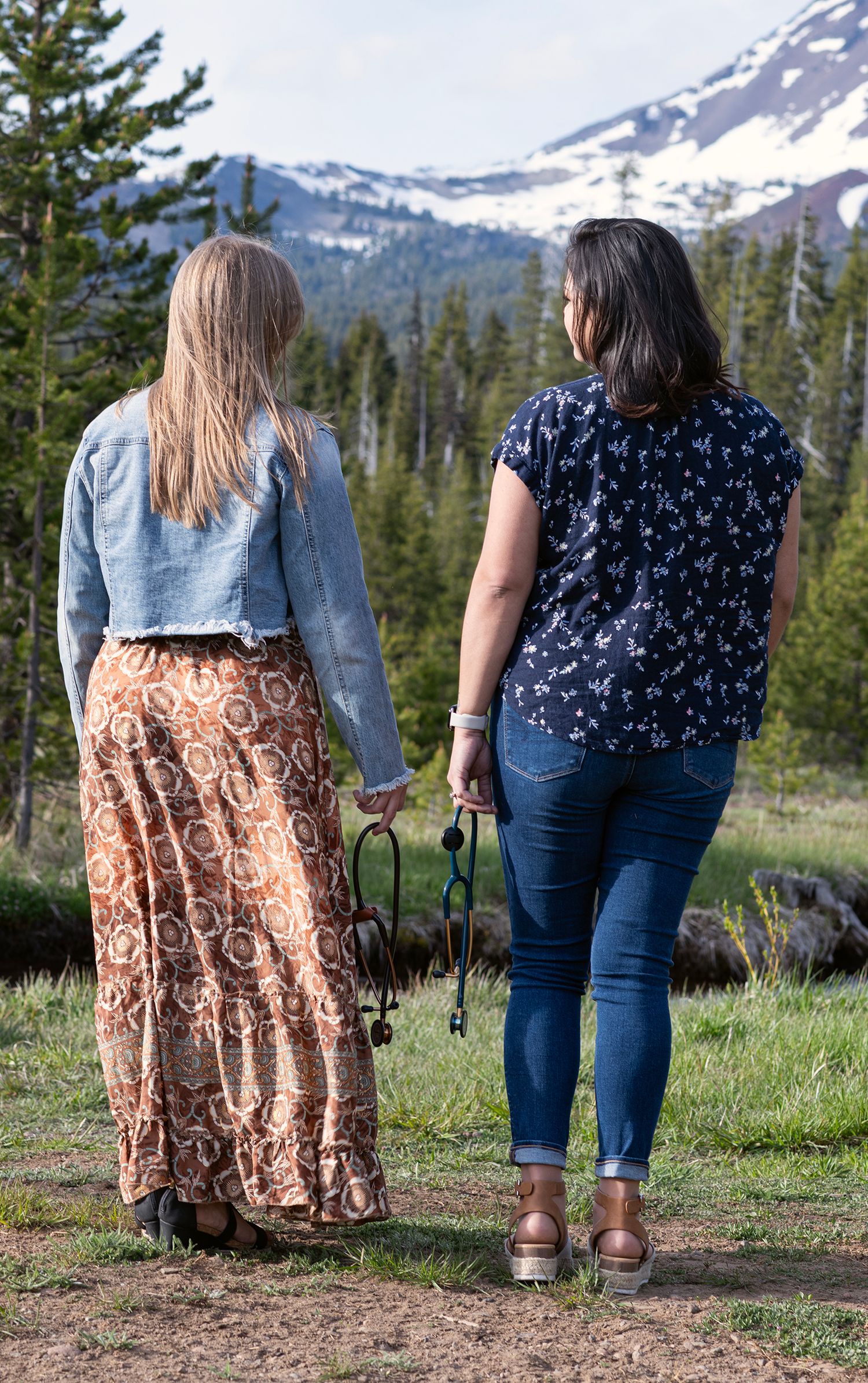 Two women walk hand-in-hand toward a scenic mountain landscape with snow-capped peaks and evergreen trees.